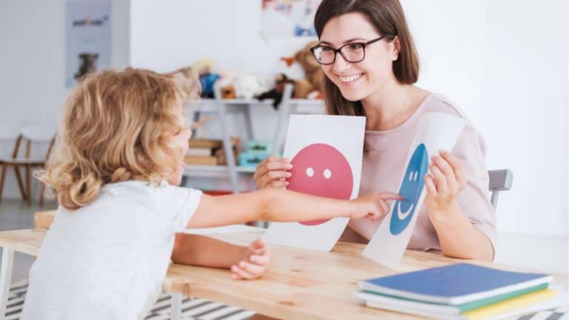 Therapist shows emotion cards to children who point at a happy face, classroom table, Activities for Autistic Kids.