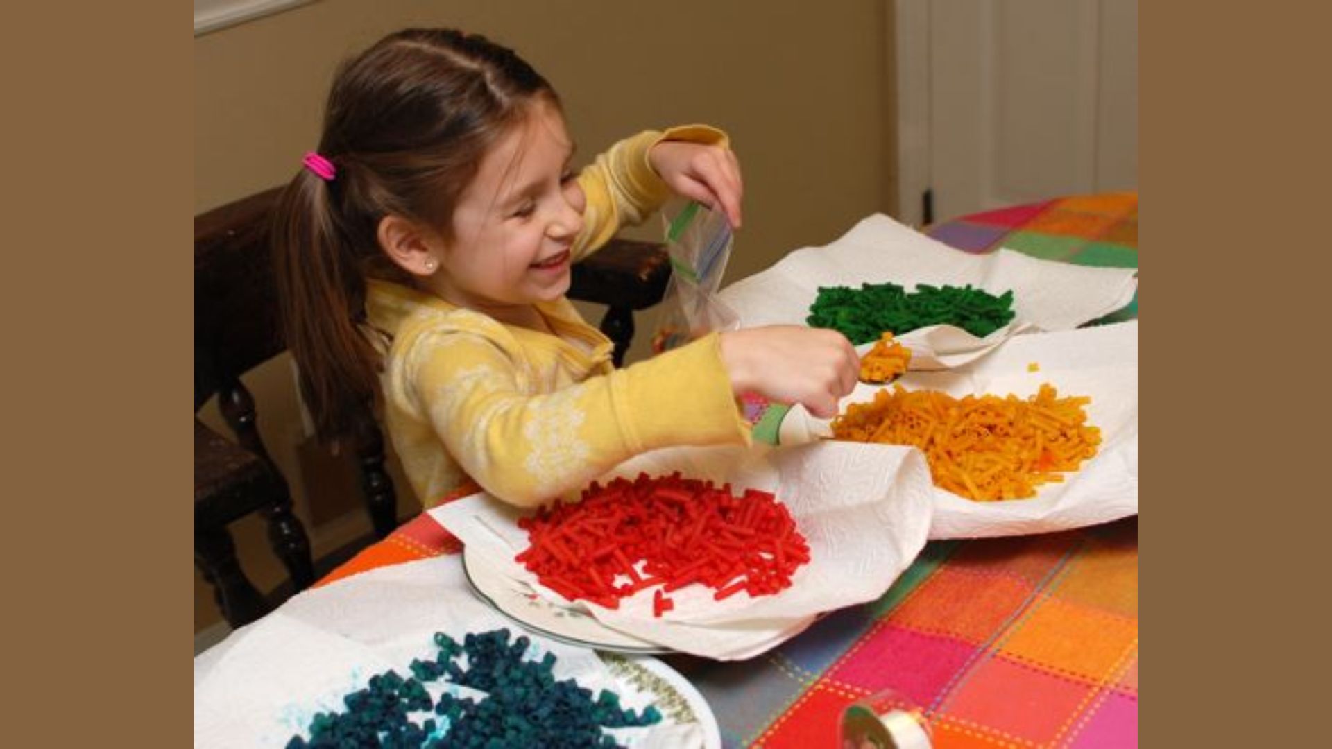 Smiling preschool girl sorting red, yellow, green, and blue dyed pasta into bags, practicing fine motor skills and sharing materials in a friendship activity for kids