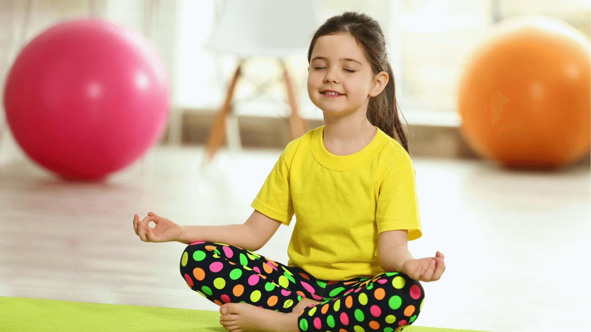 Smiling girl in yellow shirt meditates on green mat, colorful exercise balls behind, Activities for Autistic Kids.