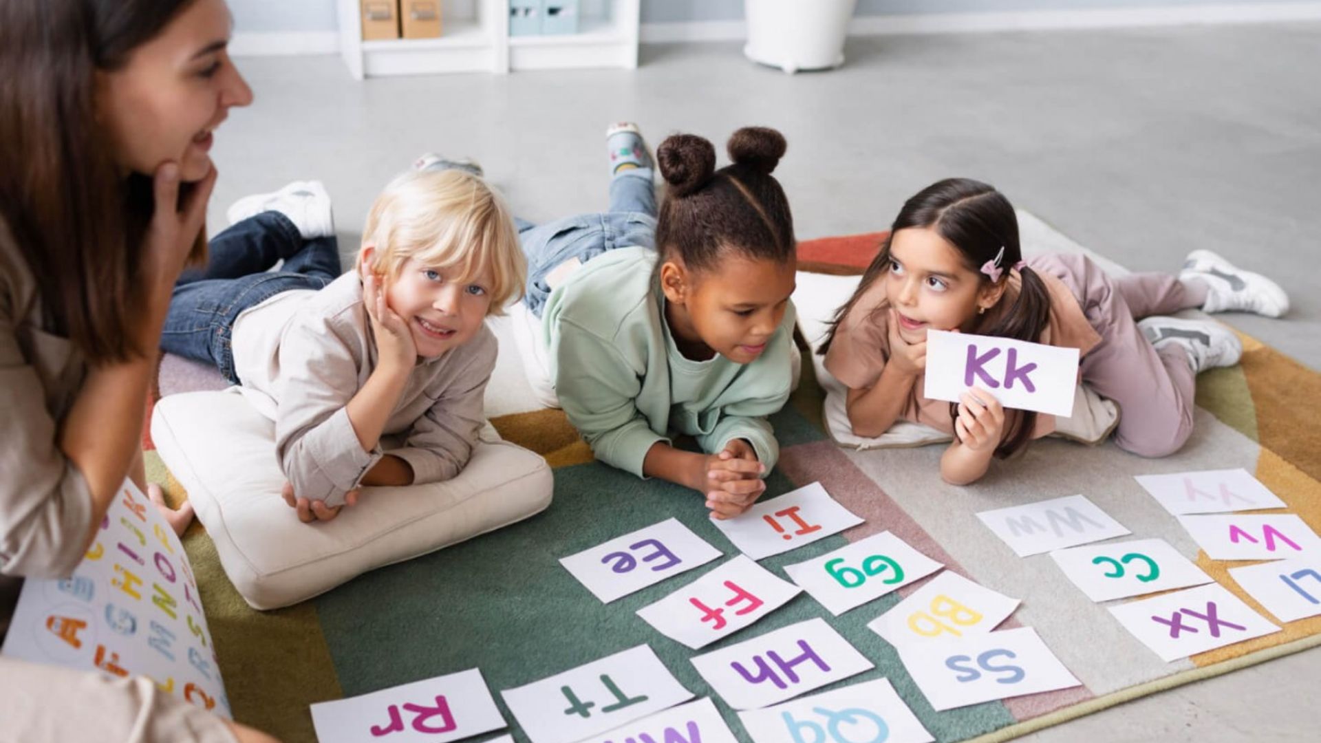 Three children lie on a colorful rug during a letter learning activity.