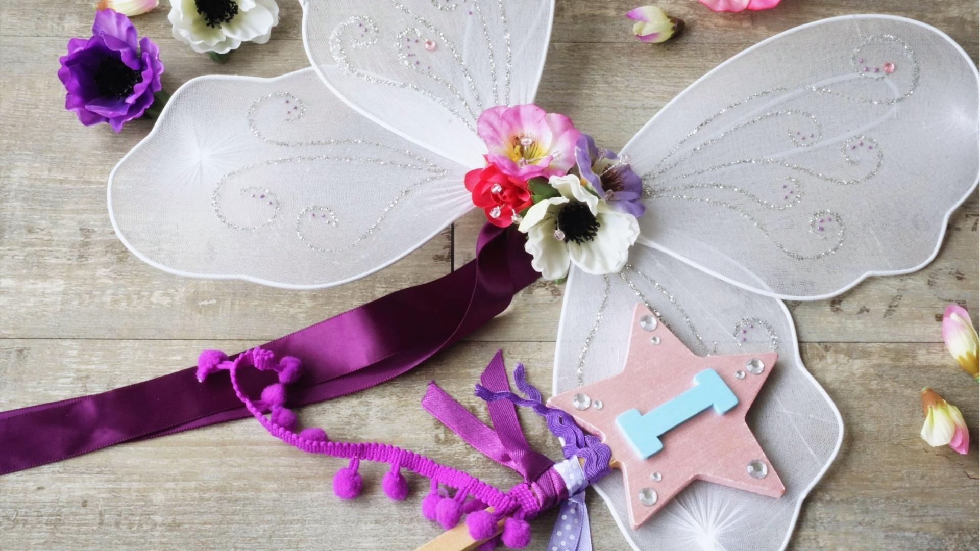 White fairy wings with flowers, purple ribbons, star wand, wood table, flowers.