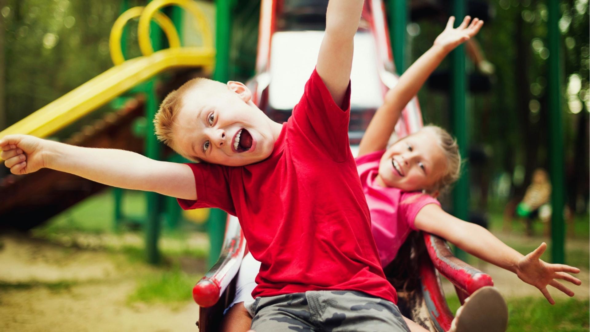 Two happy kids with arms out wide ride playground merry go round, laughing, sunny, Summer Activities for Kids.