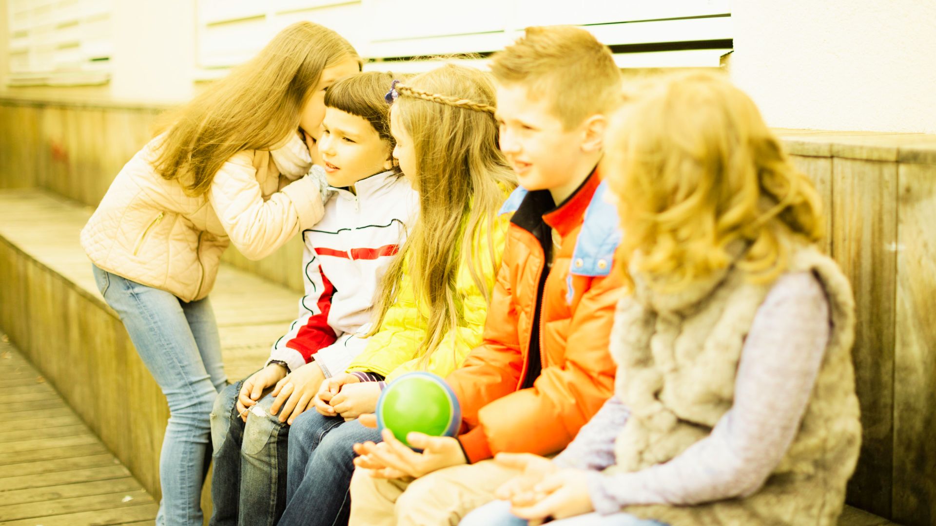 A group of children sitting together on a wooden bench in a park, smiling and chatting with each other.