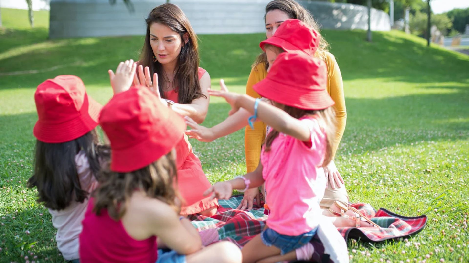 Preschool children in red hats playing a group hand-clapping game with teachers on grass, building cooperation and social rhythm through an outdoor friendship activity