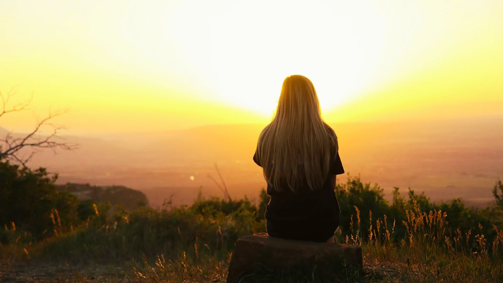 A woman sitting on a rock, gazing at the sunset, with vibrant orange and purple hues in the sky.