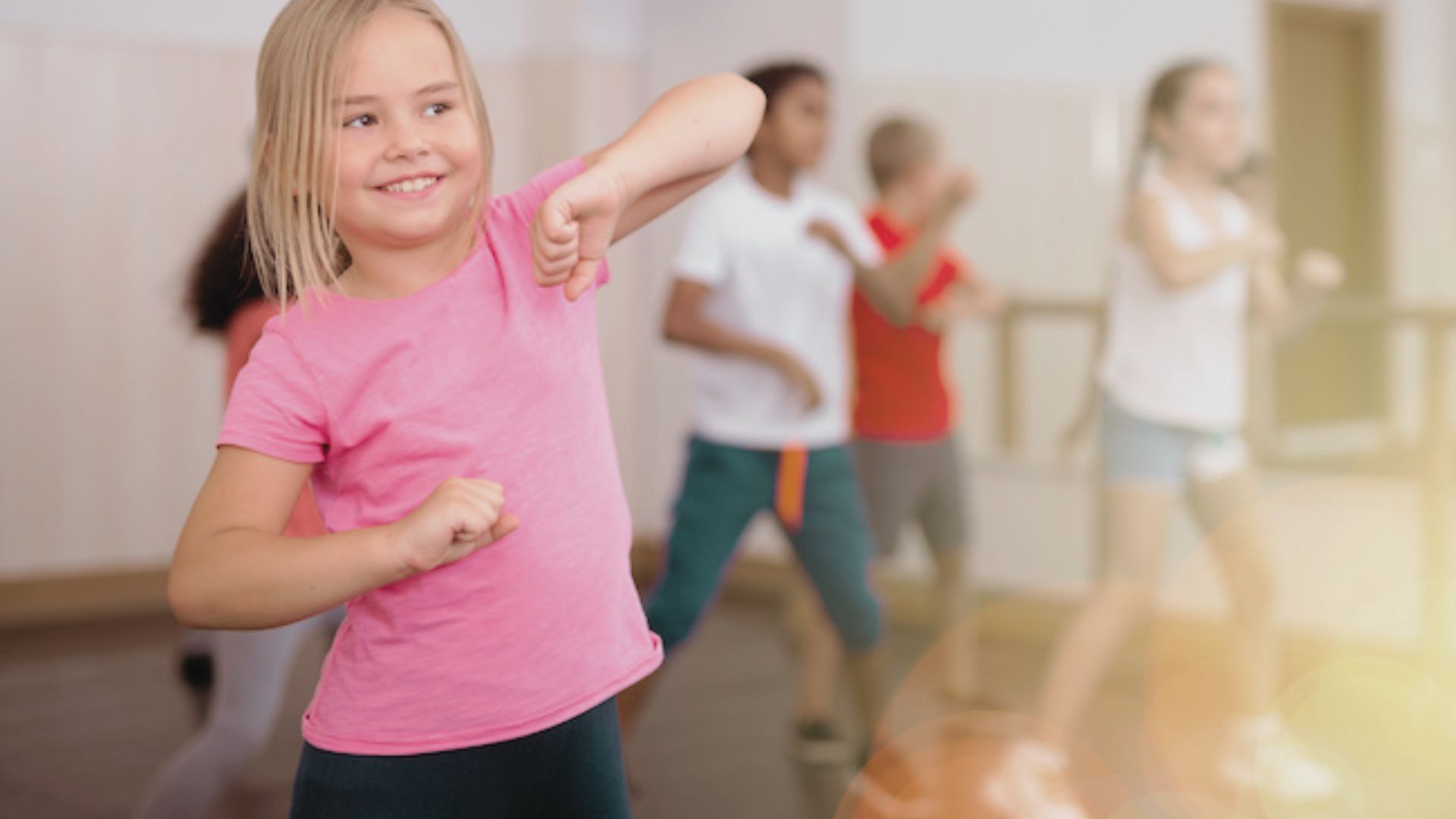 A girl in a pink shirt smiles and dances with her arm raised, while other kids follow in the background.