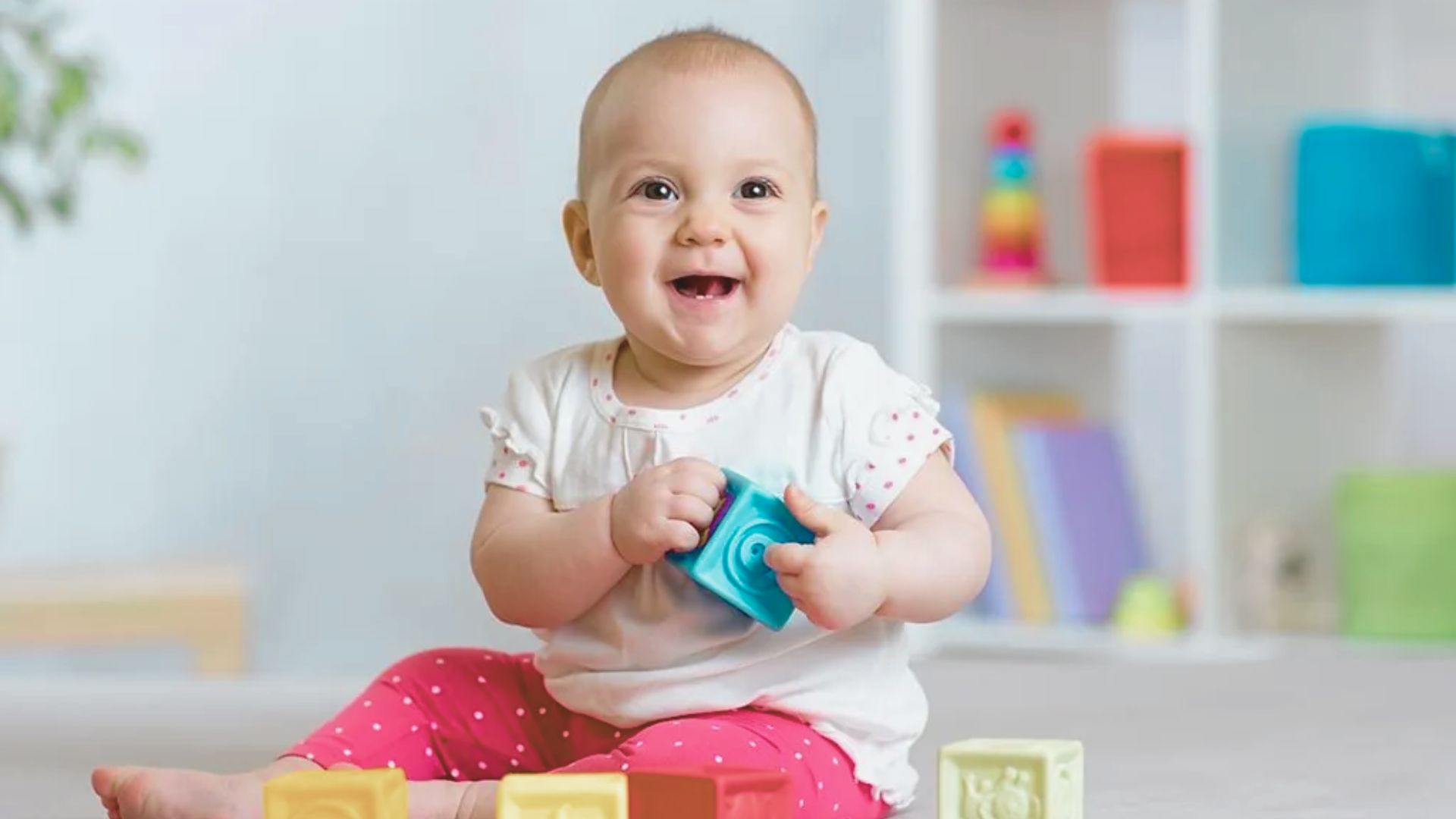 A happy baby in a white top and pink polka-dot pants sits with colorful blocks, holding a blue toy.