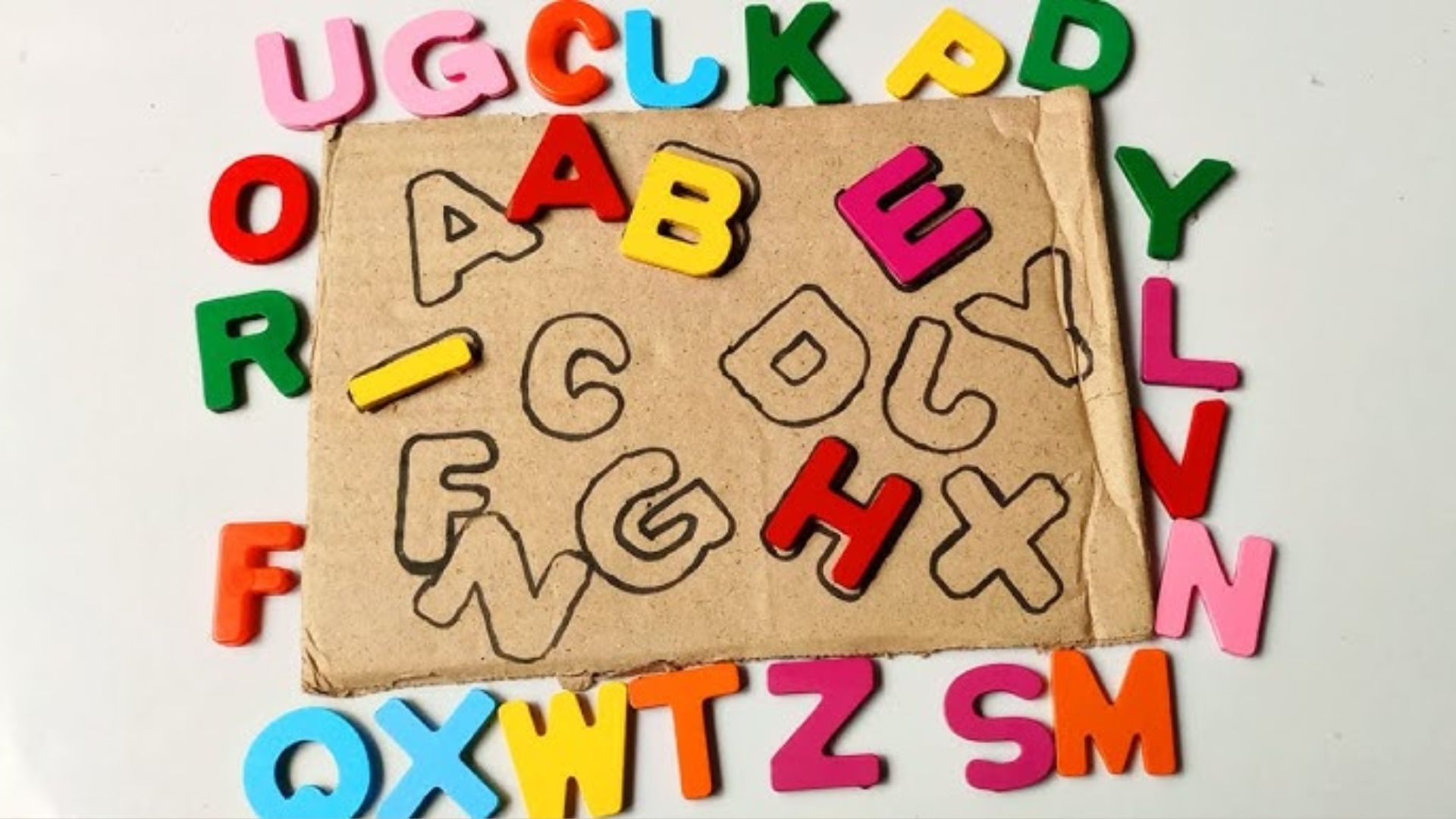 Cardboard square with hand-drawn letter outlines A, B, C, D, F, G, H, X. Colorful plastic letters A, B, E, H placed on matches.