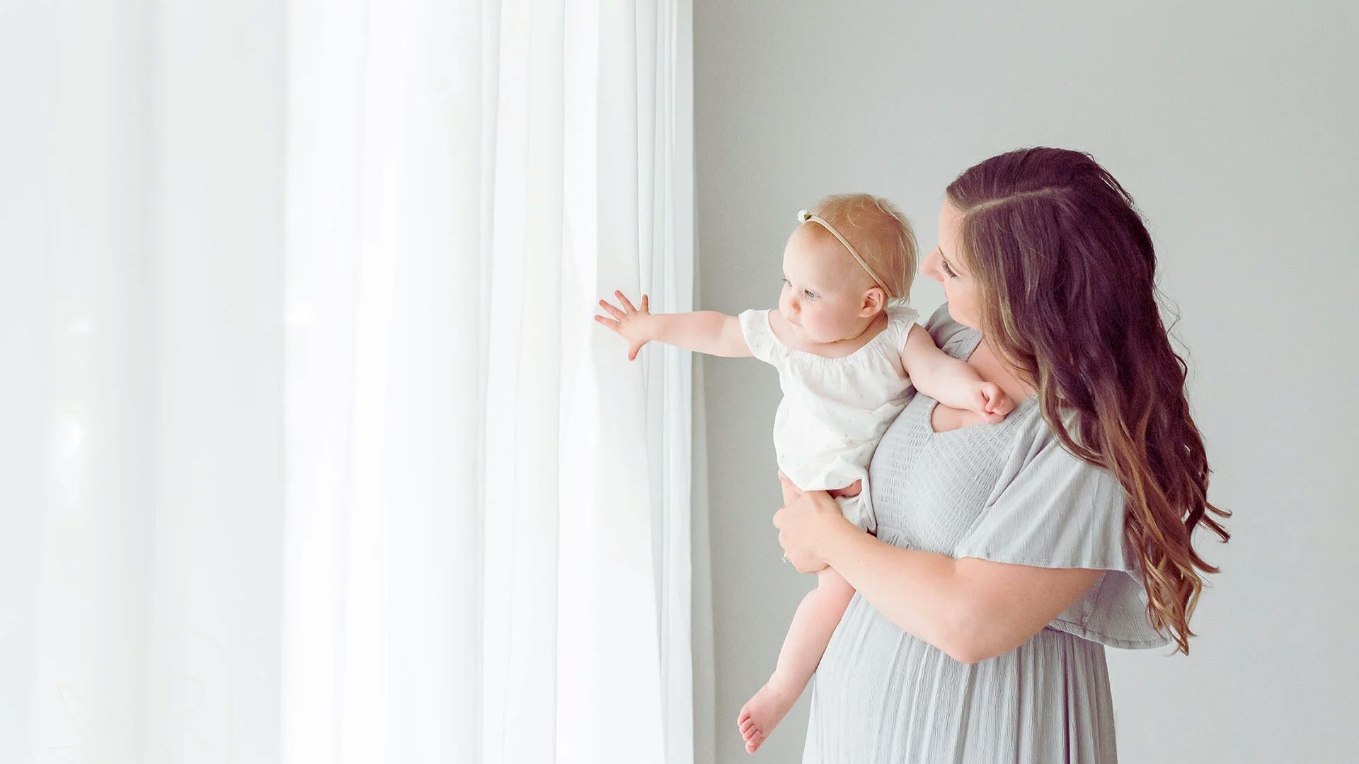 . A mother holds her baby close while standing by a window, highlighting their bond in a serene setting.