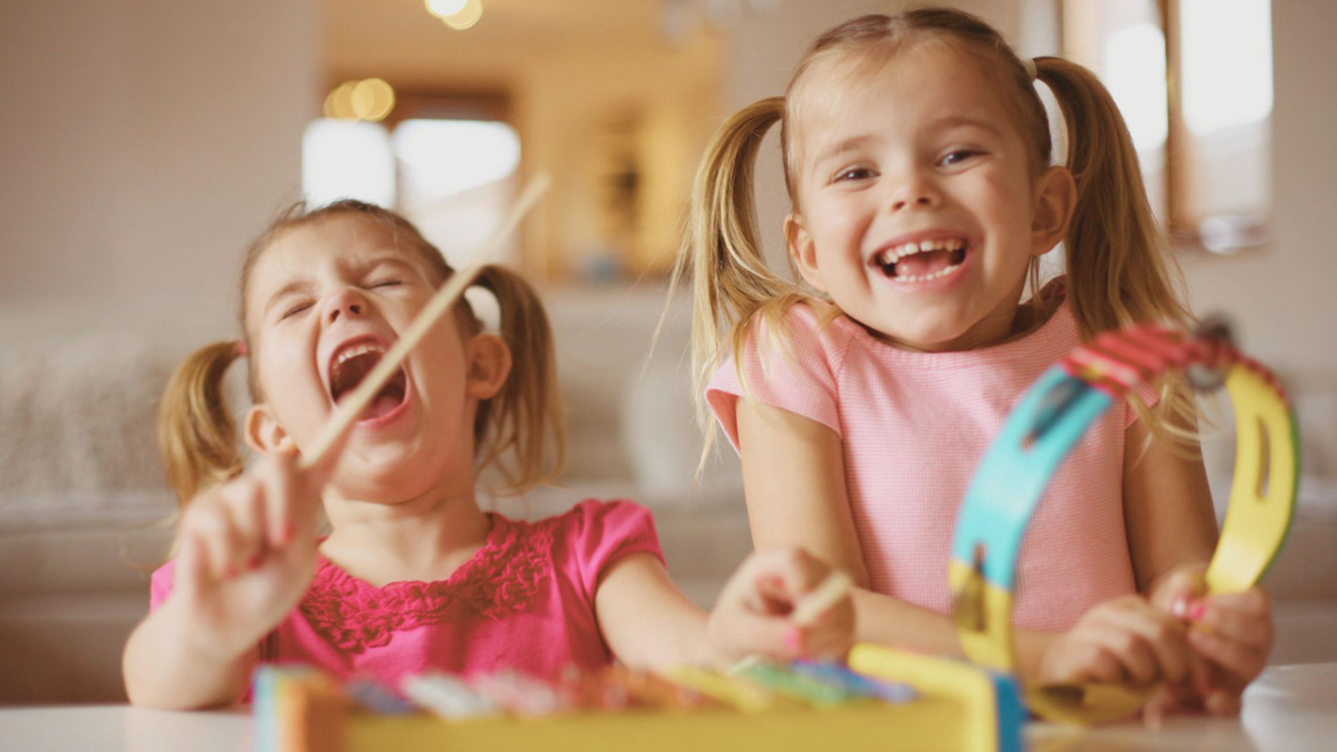 Two young girls in pink shirts laugh while one plays a tambourine and the other taps a rhythm stick.