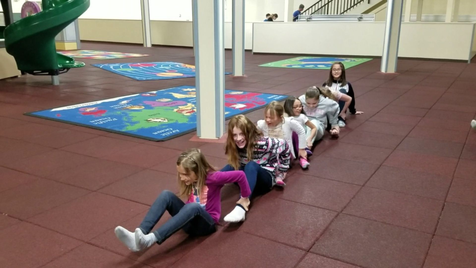 Kids participating in a group activity indoors, sitting in a line and holding on to each other while smiling and playing together.