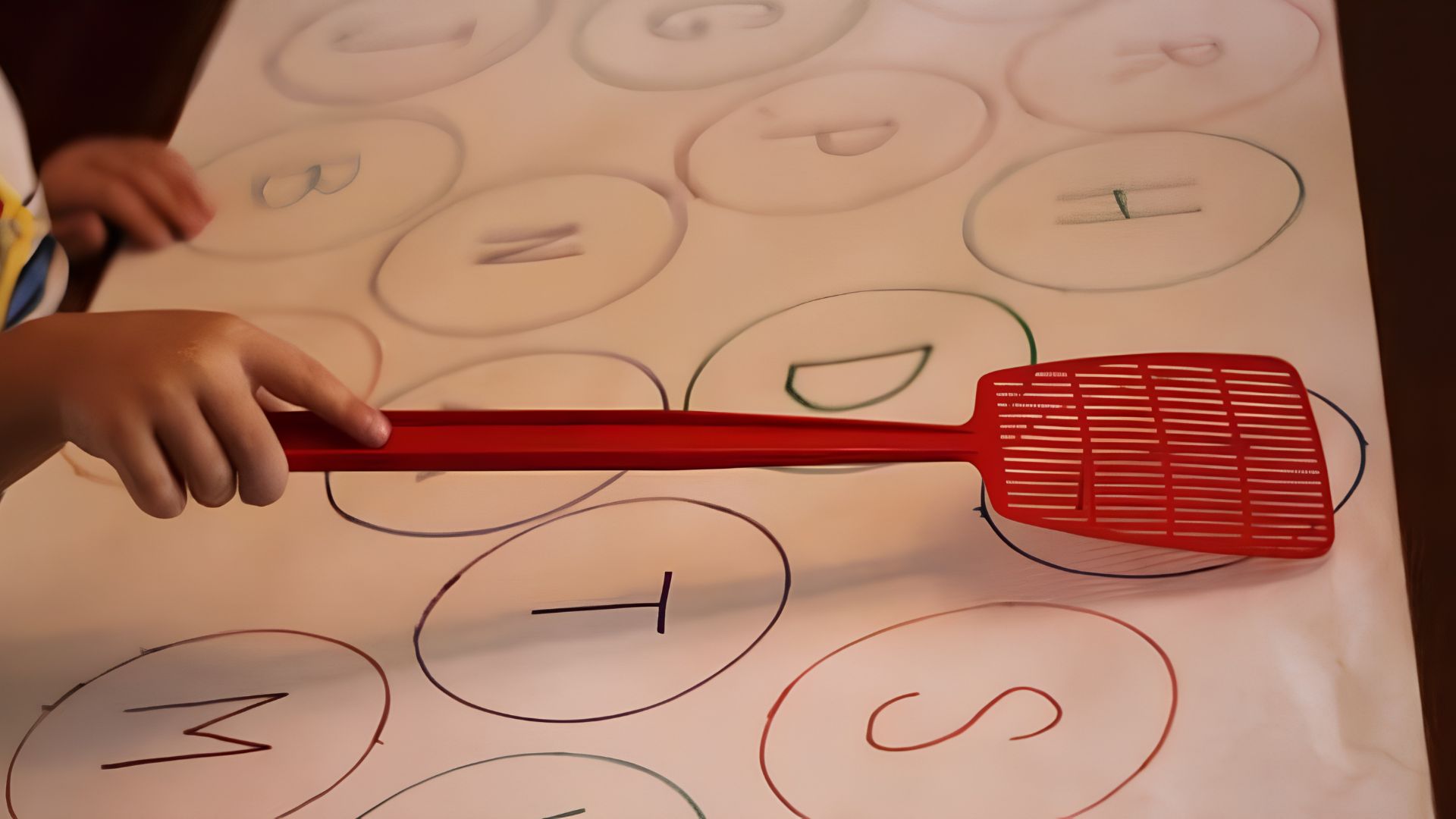 Child’s hand holding red fly swatter over large paper with hand-drawn letters in circles, ready to swat called letters.