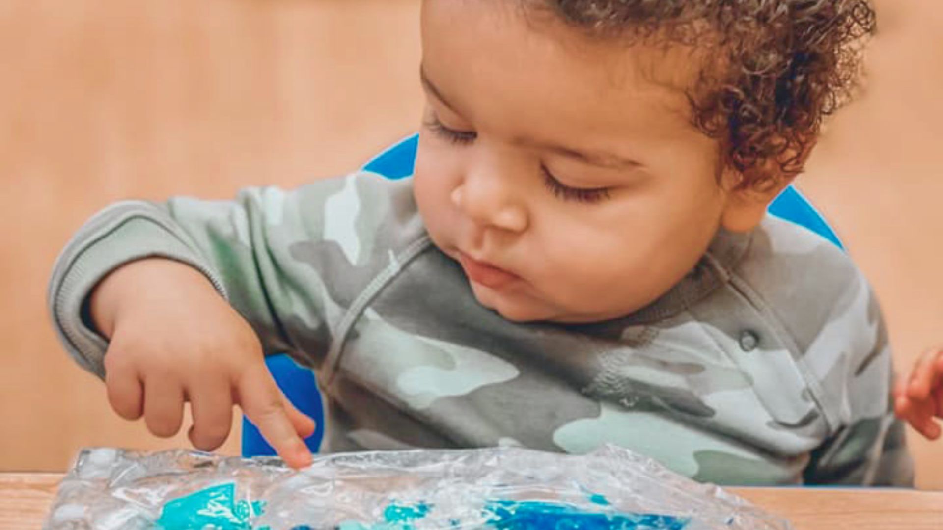 A curly-haired infant in a camouflage shirt presses a finger into a sealed sensory bag filled with blue gel.