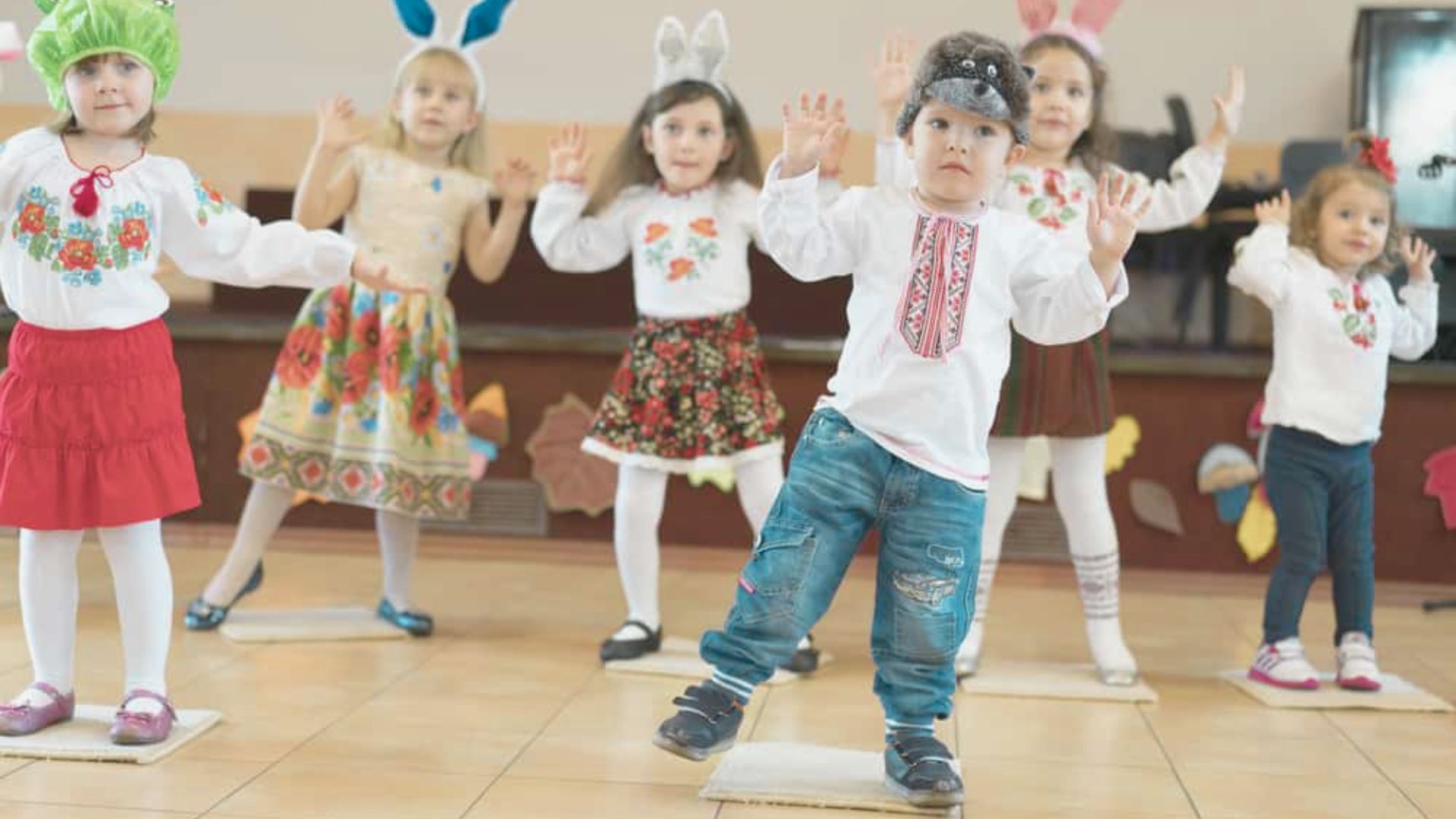 A group of young children wearing colorful outfits and animal headbands dance together on small mats.