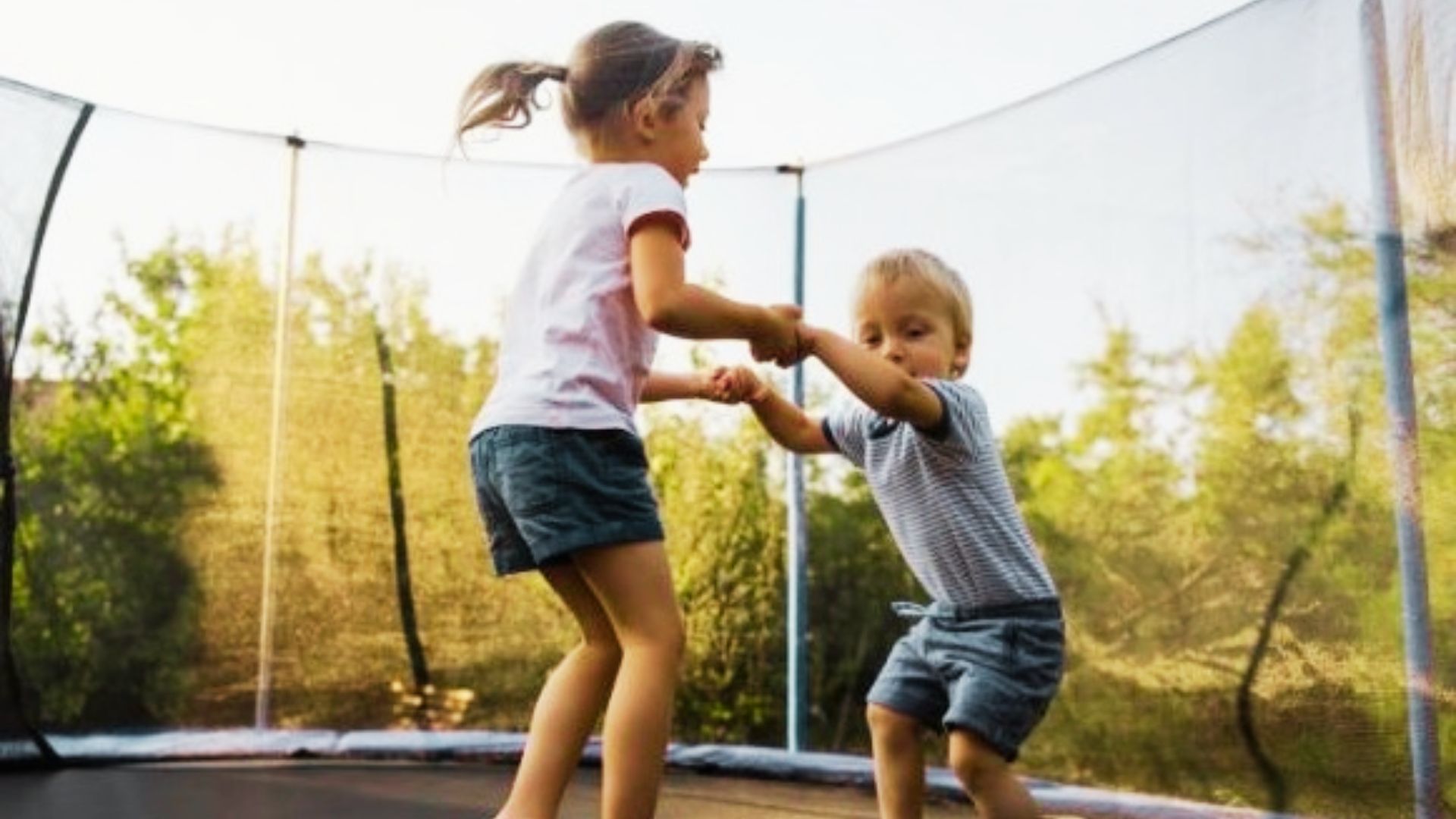 Two children joyfully jumping on a trampoline in a sunny backyard, surrounded by grass and trees.