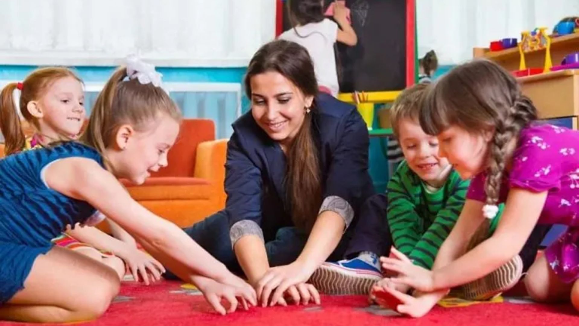 Smiling teacher sitting on a red carpet with four kids, playing a hands-on group activity game in a colorful classroom at school