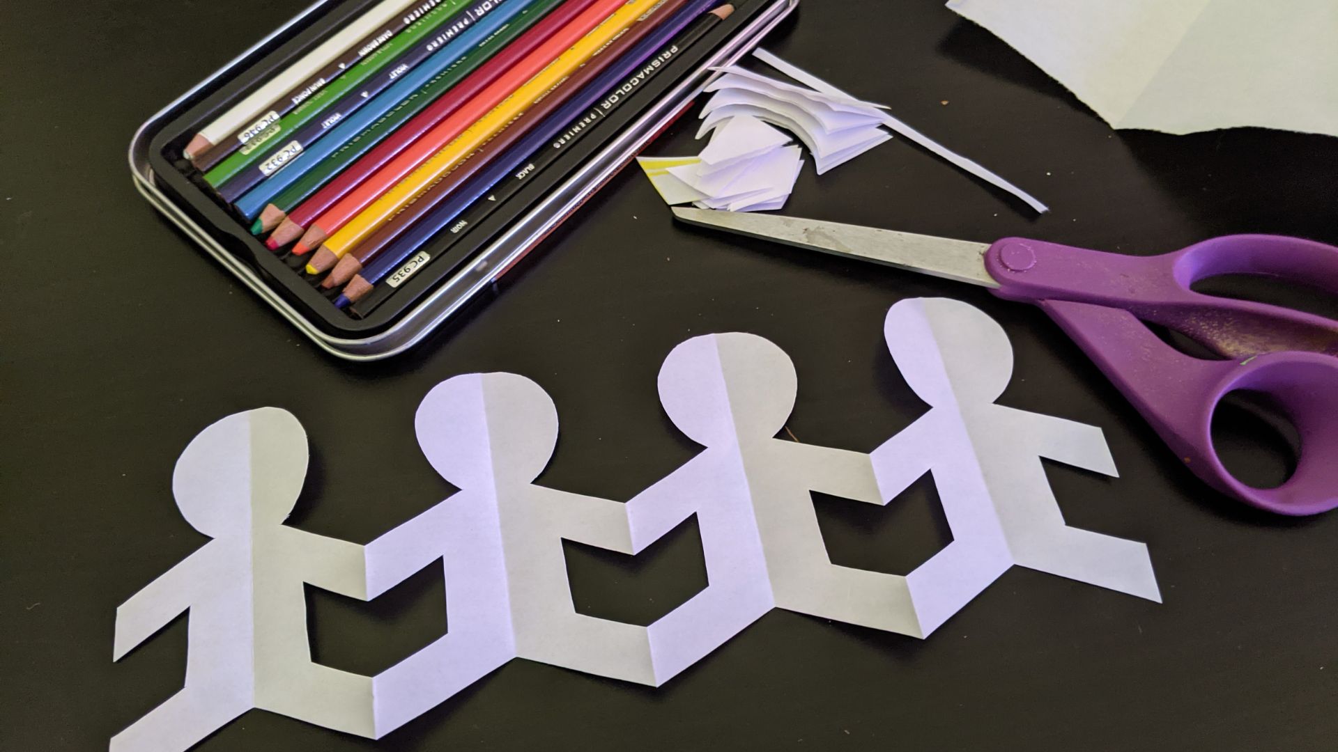 White paper chain of connected people cutouts next to scissors and colored pencils, symbolizing unity and teamwork in a friendship craft activity for kids