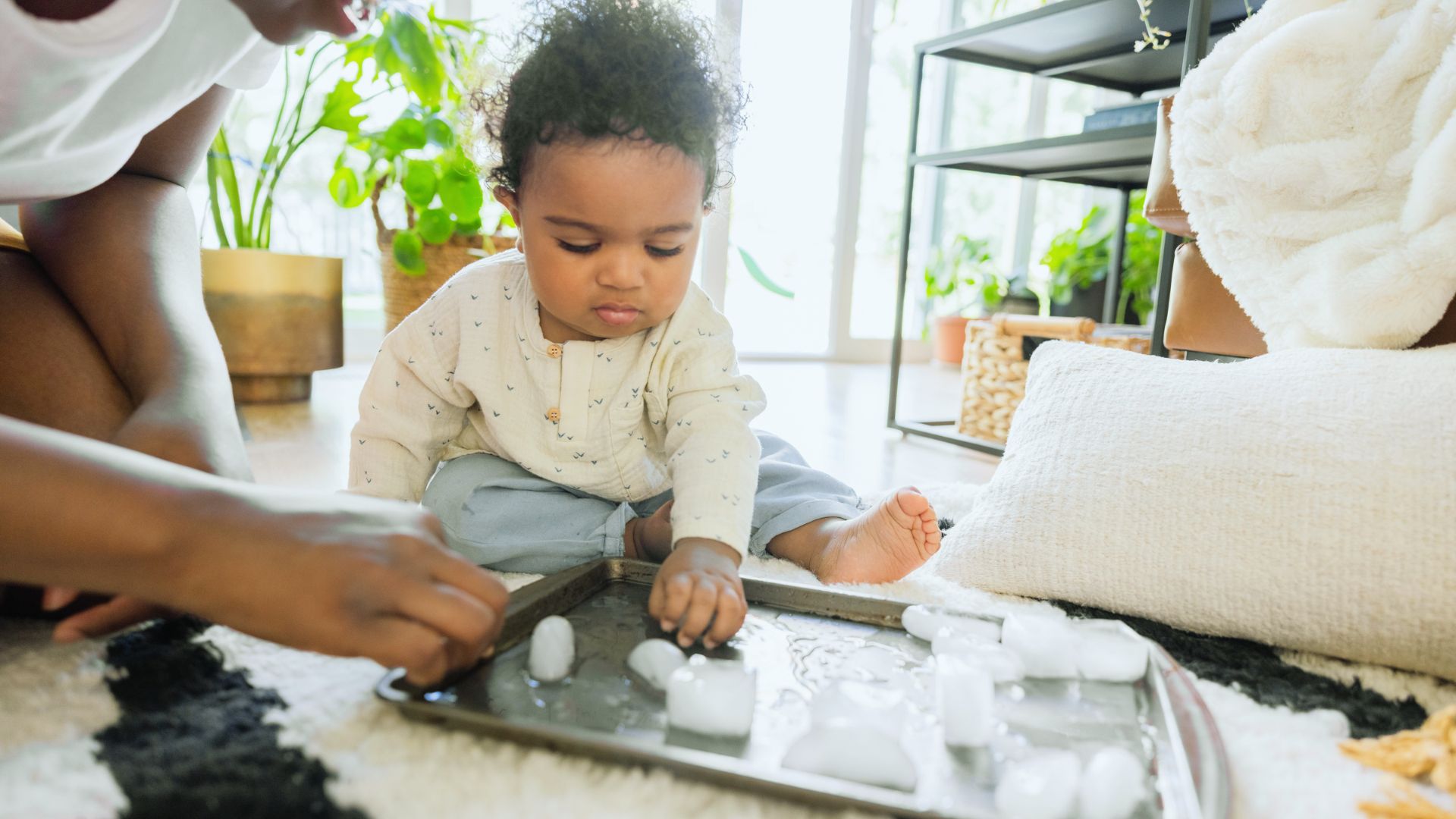A curly-haired infant sits on a rug reaching for ice cubes on a metal tray with an adult nearby for guidance.