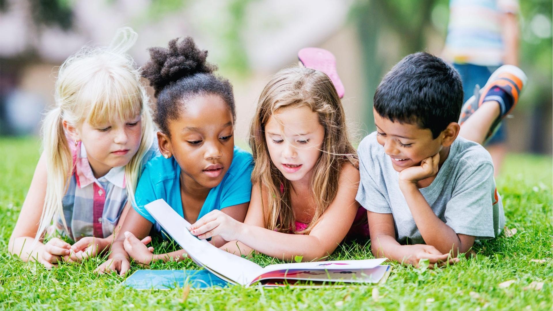 Four diverse kids lie on grass reading a book together outdoors, smiling, sunshine, Summer Activities for Kids.