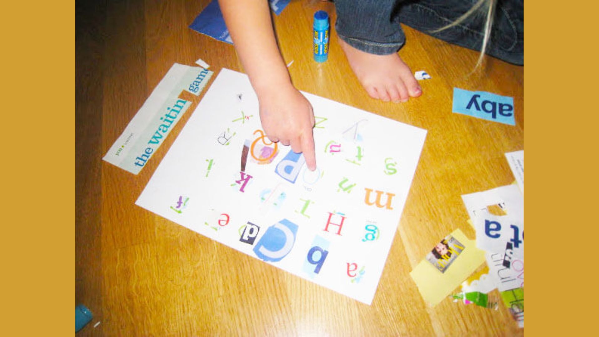 Child’s finger pointing to cut-out magazine letters glued on white paper to make an alphabet collage, with glue stick nearby.