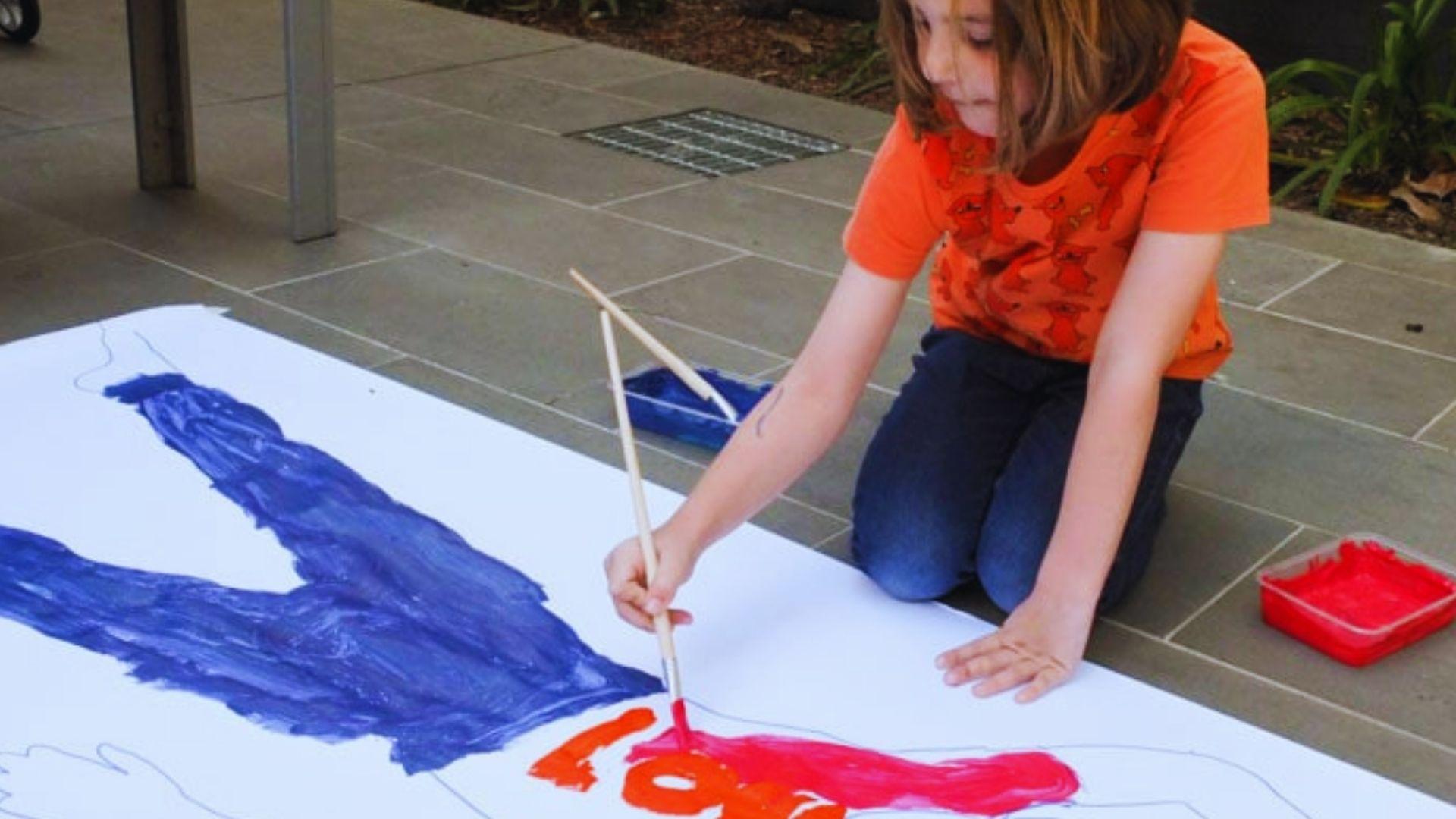 A young girl joyfully painting on a large sheet of paper, surrounded by colorful art supplies.