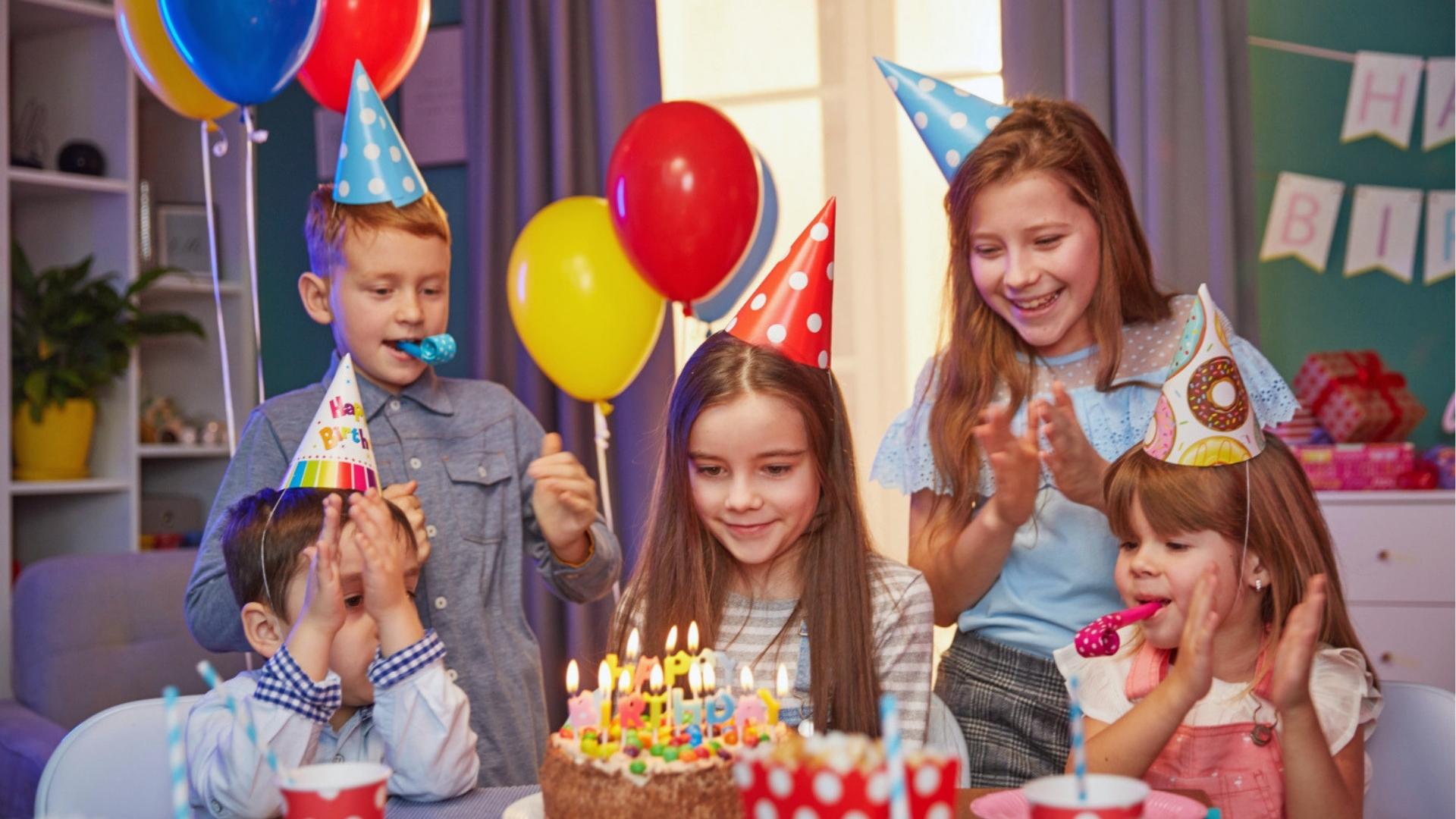 Kids wearing party hats around birthday cake with candles, balloons, blowing noisemakers.