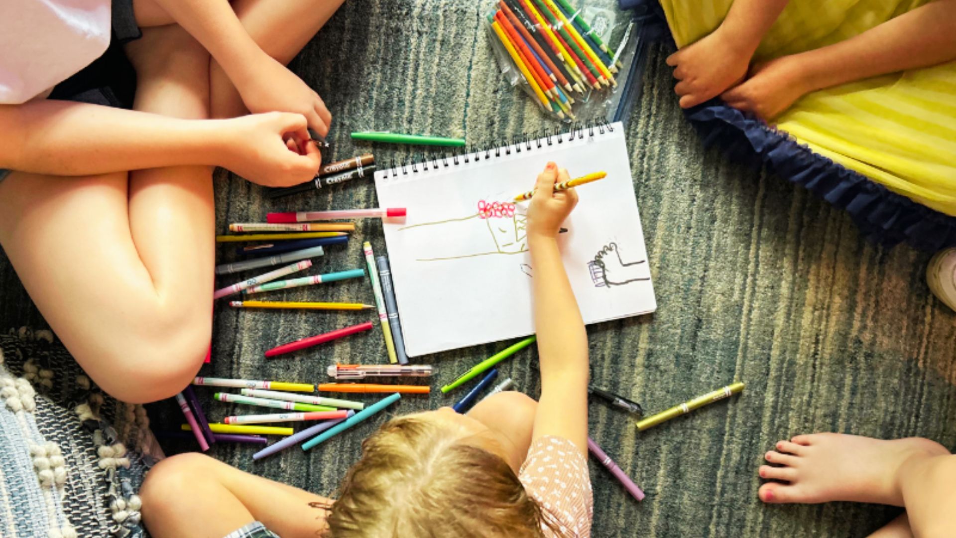 Children using colorful markers to draw on a large piece of paper spread out on a table.