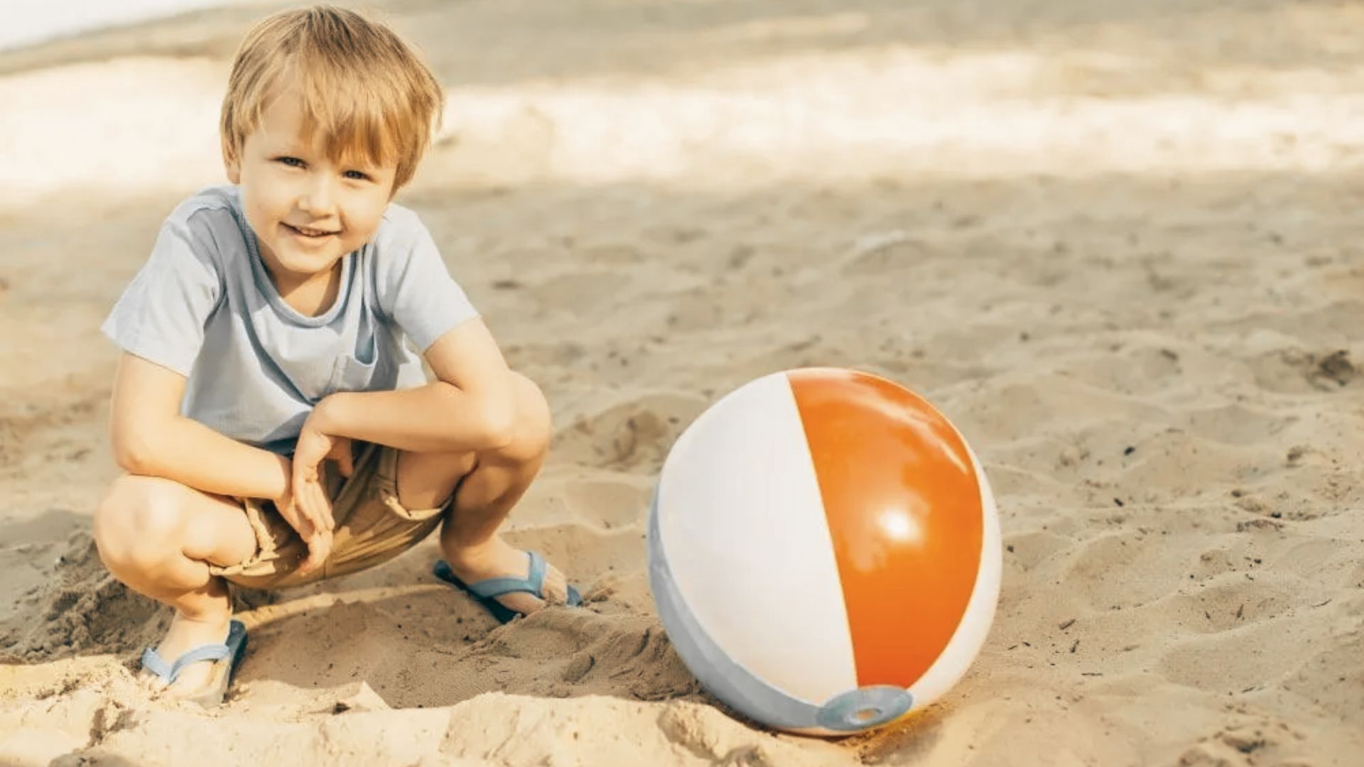 A young boy sits on the sand, smiling while playing with a colorful beach ball beside him.
