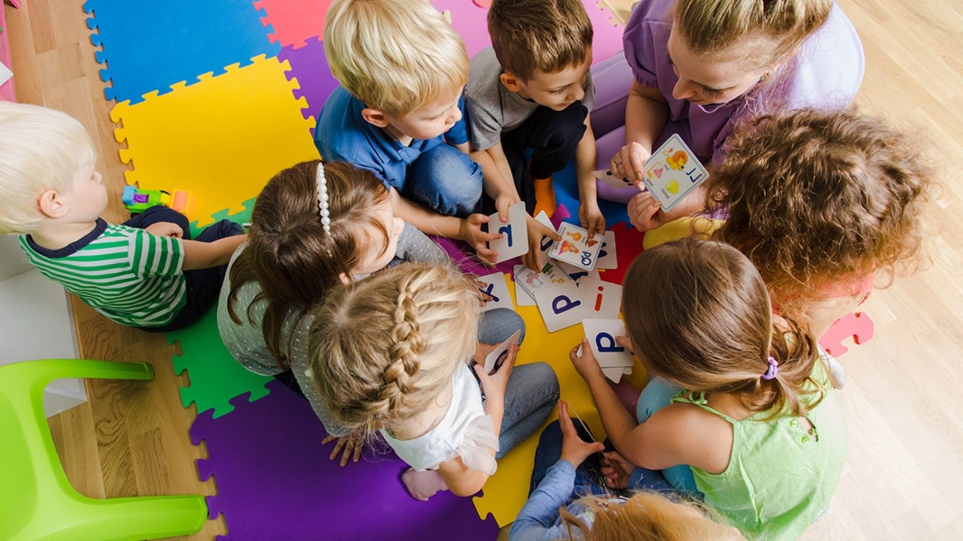 Overhead view of teacher and six preschool kids sitting on foam mats, playing an alphabet matching card game as a group literacy activity at school