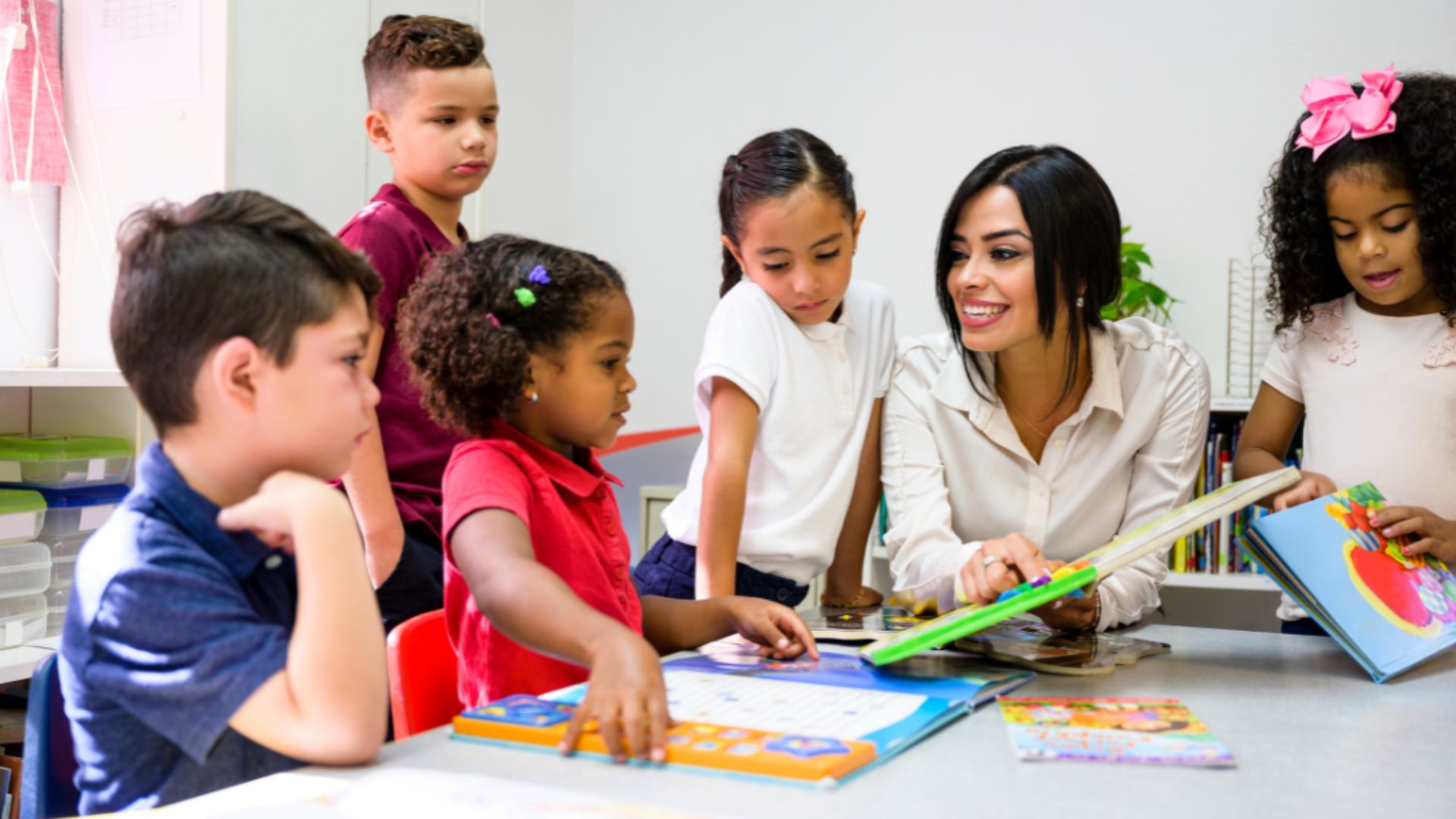 A teacher smiles while reading a picture book with five diverse elementary students gathered around a classroom table.