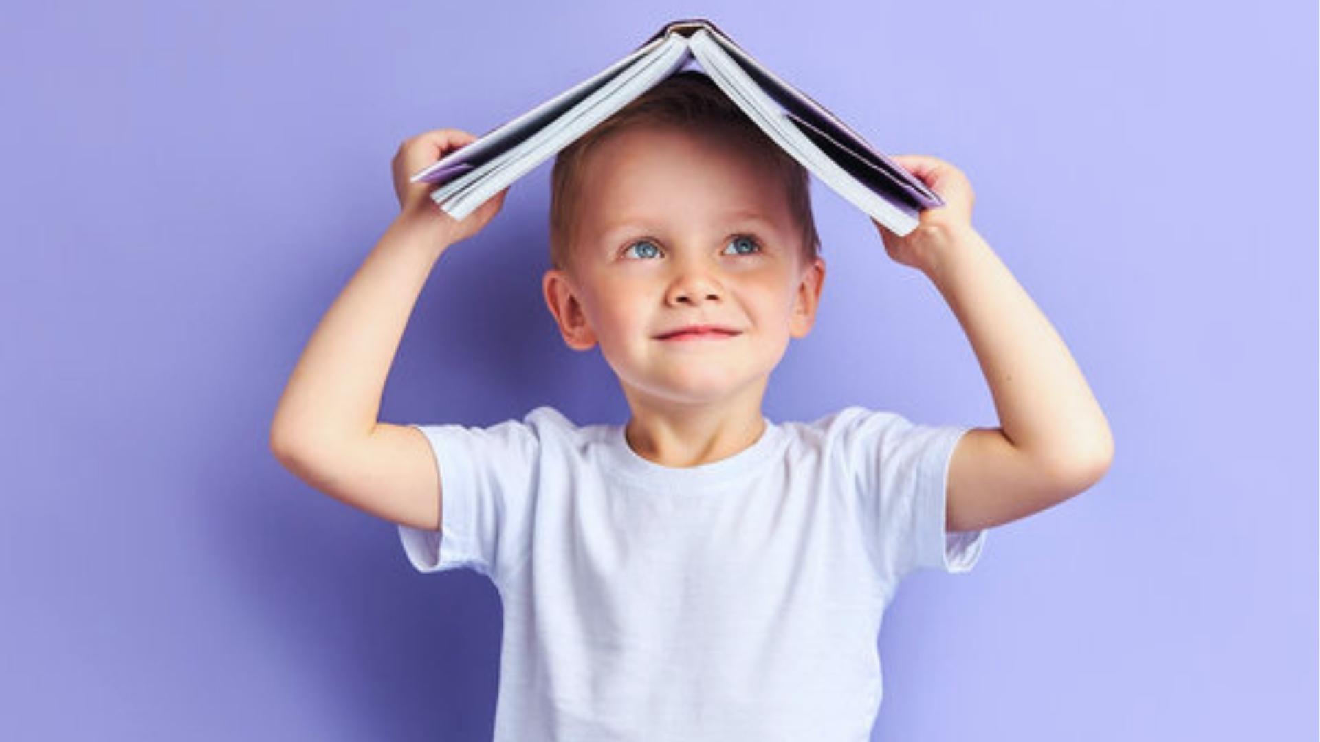 Smiling boy holds open book over head like roof, purple background, white shirt.