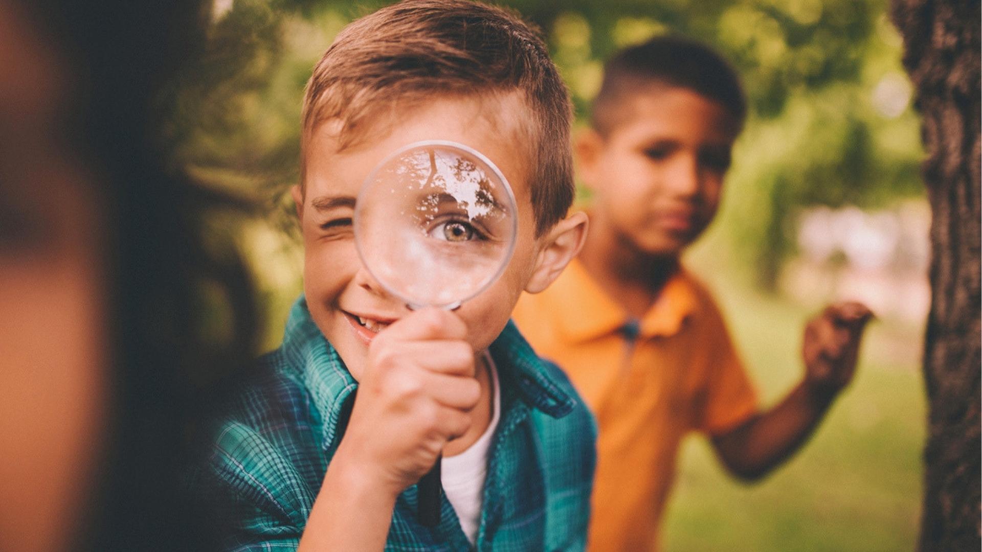 Smiling boy holds magnifying glass to eye, friend nearby, outdoor scavenger hunt, green.