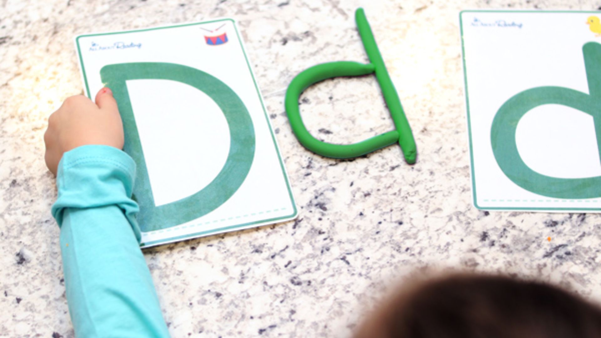 Child’s hand next to uppercase D card and green playdough shaped into lowercase d, with second D card nearby on granite counter.