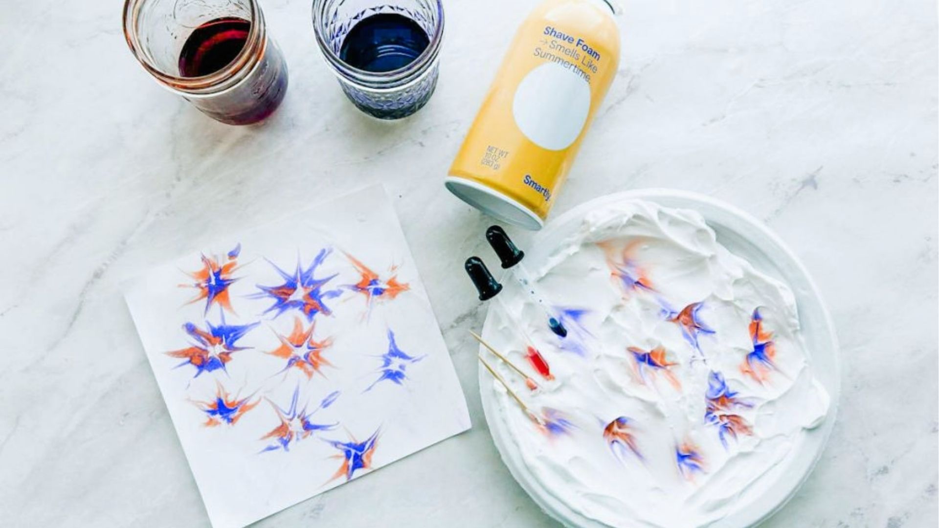 Shaving cream marbling setup with red and blue food coloring, droppers, and finished starburst prints.