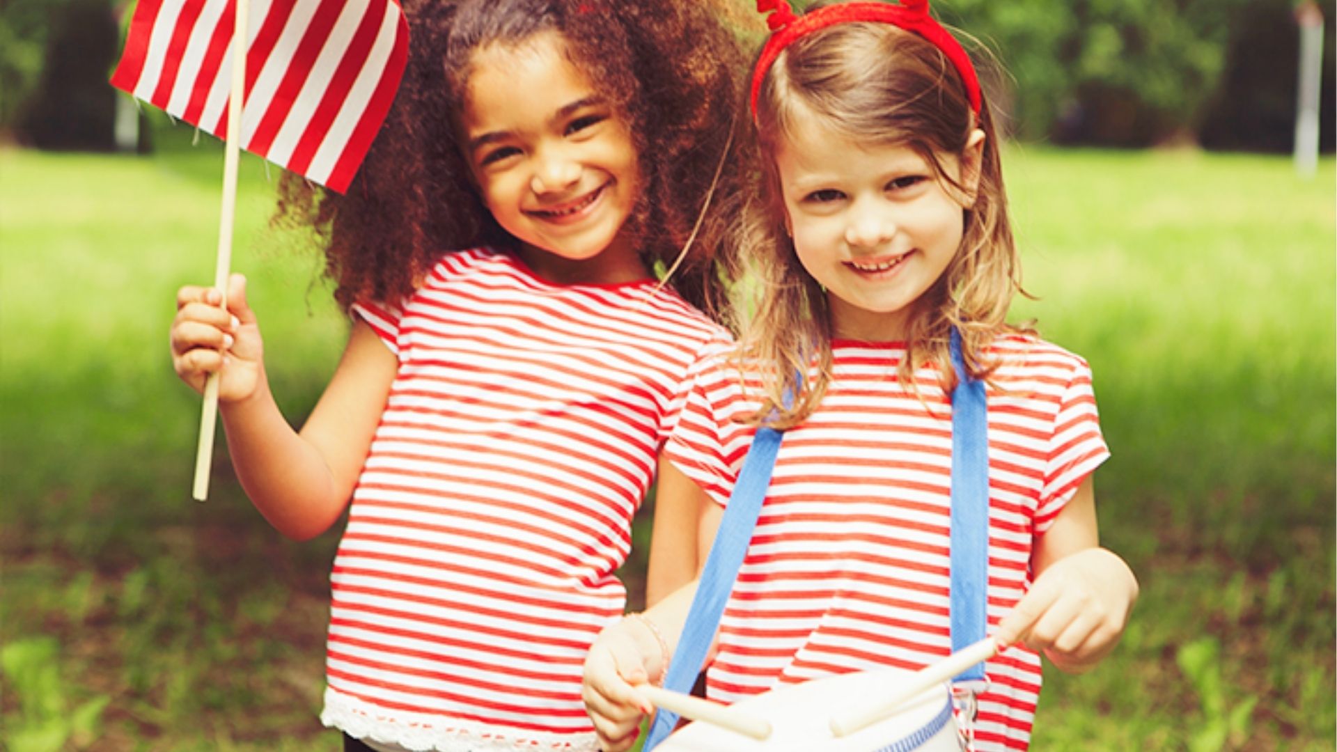Two smiling young girls in striped shirts celebrate outdoors, one holding a mini flag and one playing a drum.