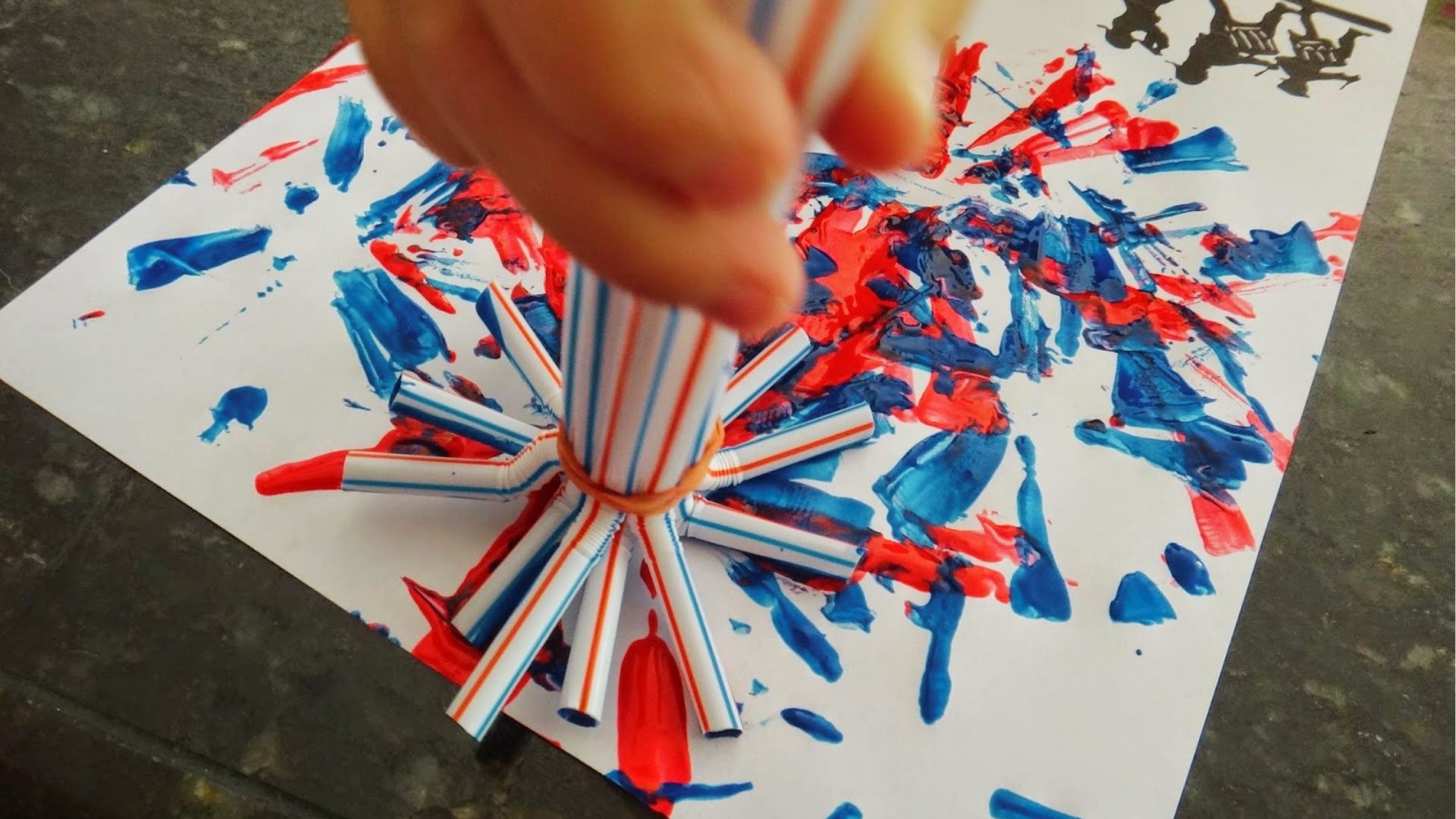 A childโs hand stamps red and blue paint with a bundle of straws to create firework bursts on paper.