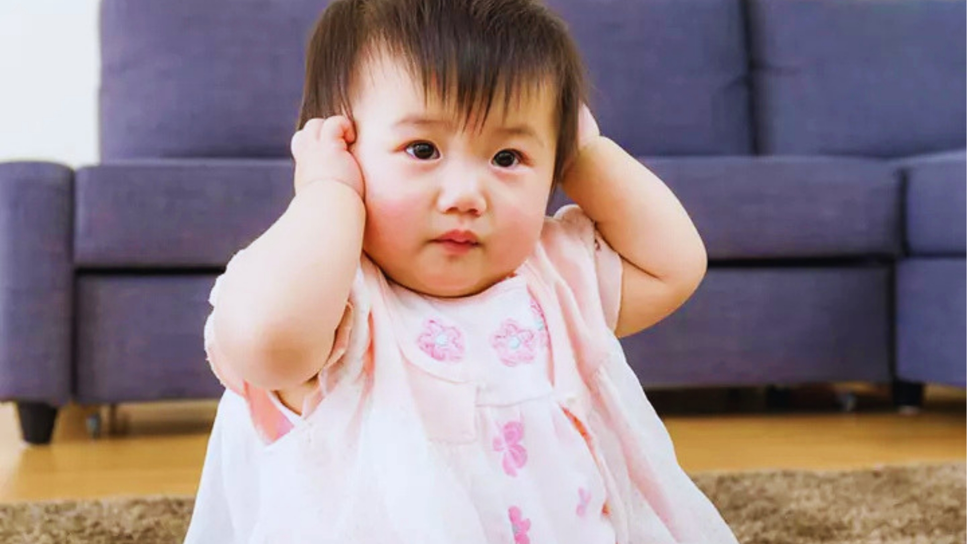 Baby in pink dress holding ears while sitting up, as head shaking sometimes accompanies ear discomfort or teething.