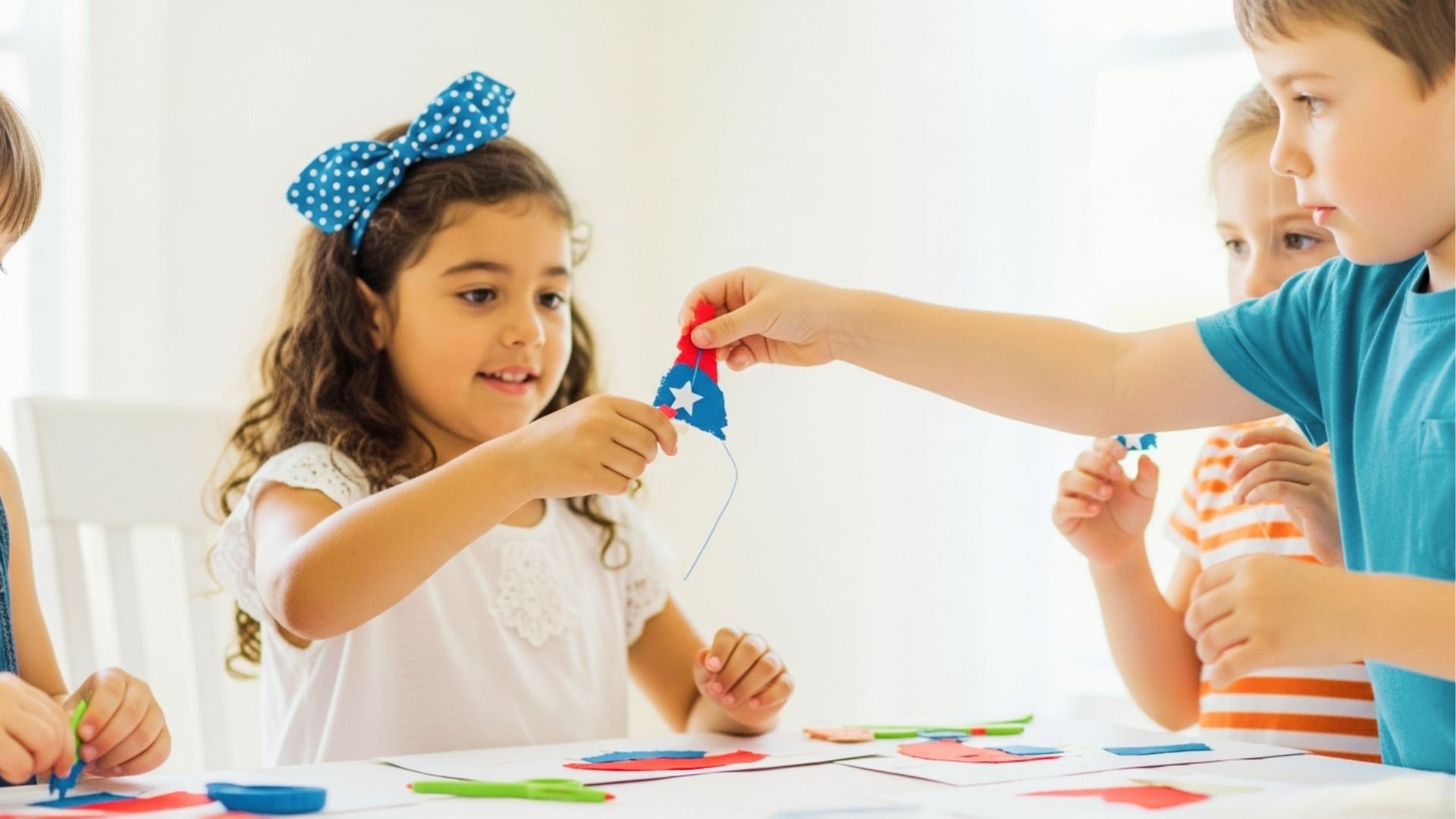 Three kids sit at a table stringing red, white, and blue felt shapes onto yarn for a craft project.