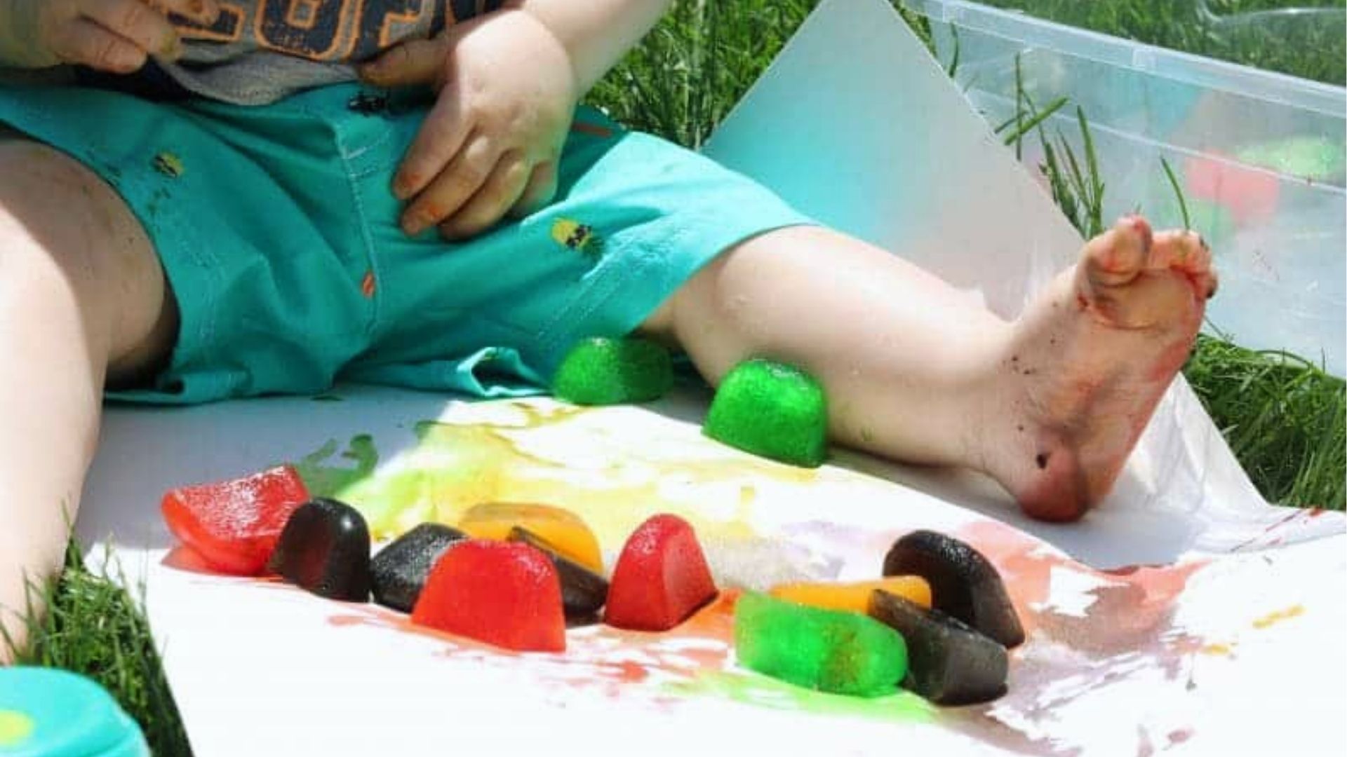 Toddler in teal shorts sits on grass painting with colorful ice cubes melting on white paper.