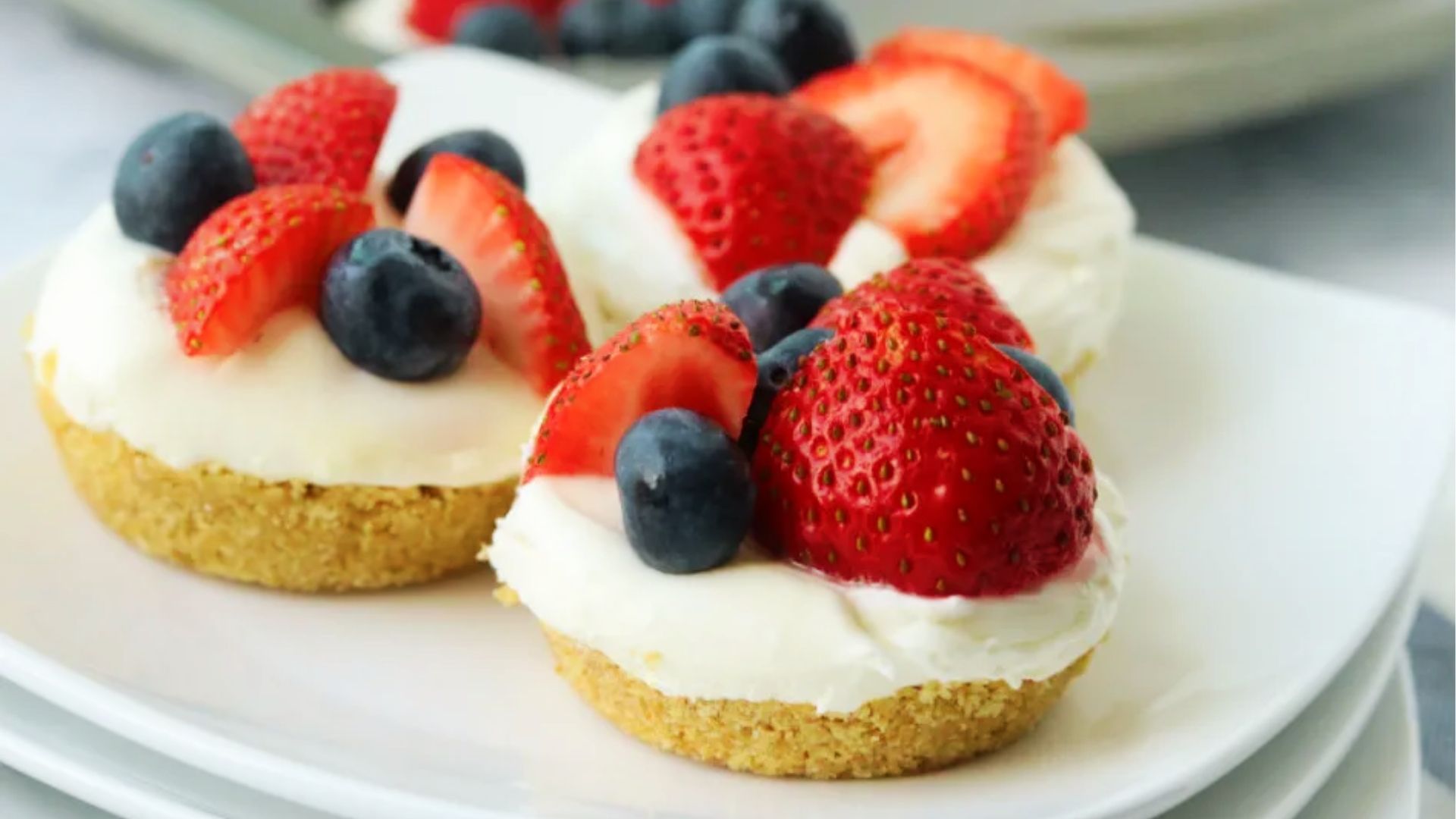 Mini cheesecake bites topped with whipped cream, strawberries, and blueberries arranged on a white plate.