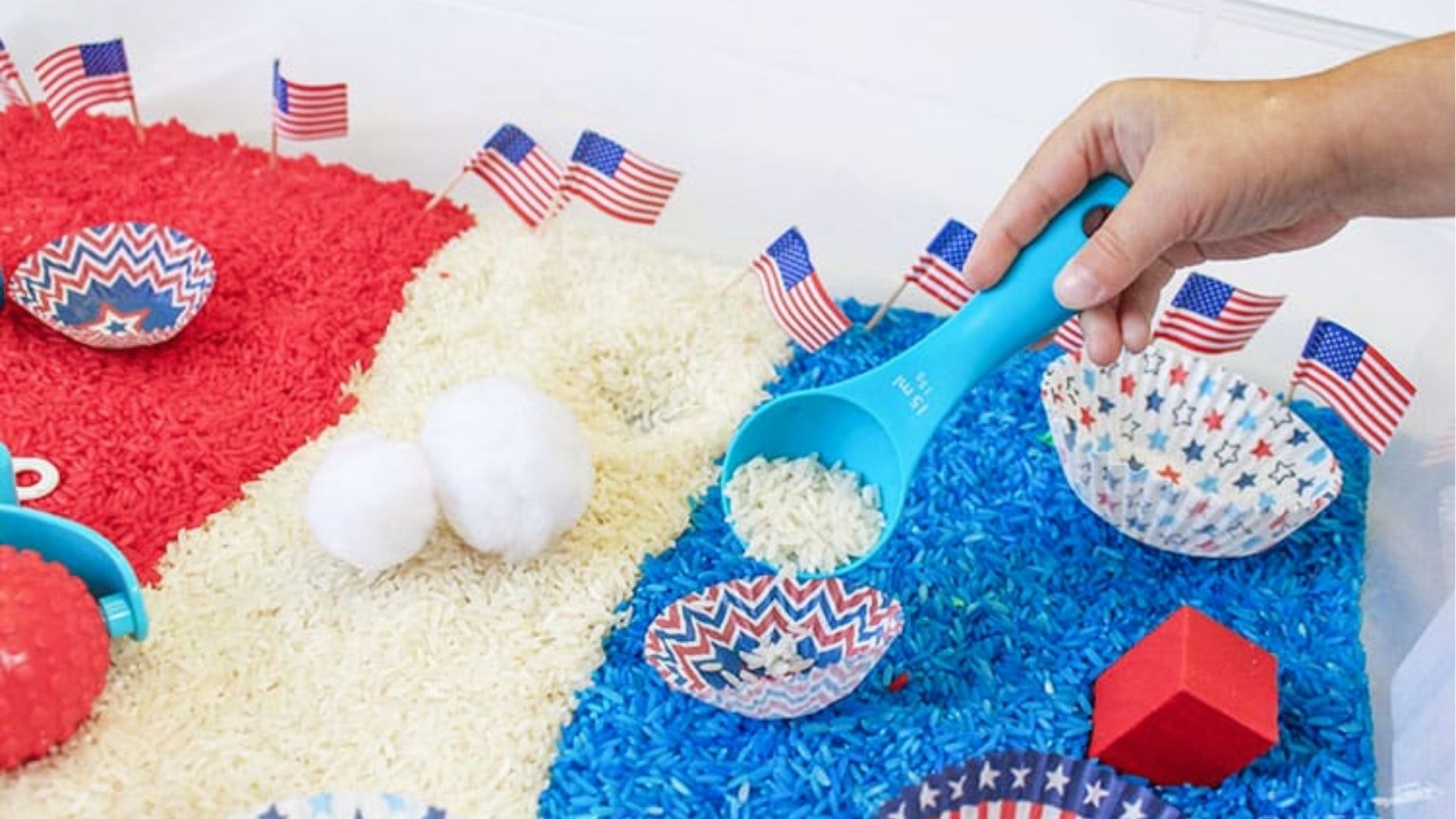Hand scoops white rice from a patriotic sensory bin filled with red, white, and blue rice, mini flags, and cups.