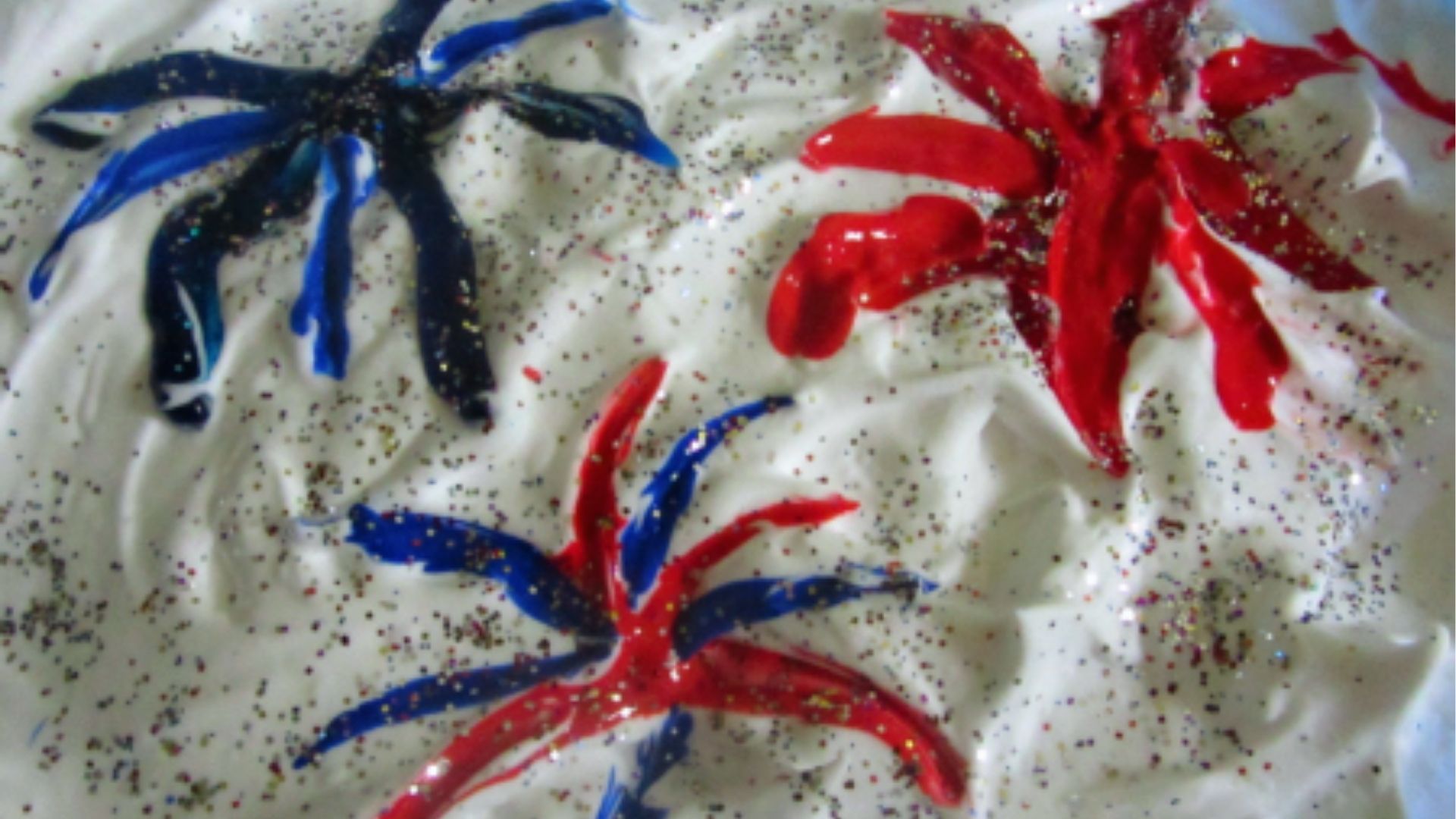 Red and blue paint streaks on shaving cream create glittery firework bursts.