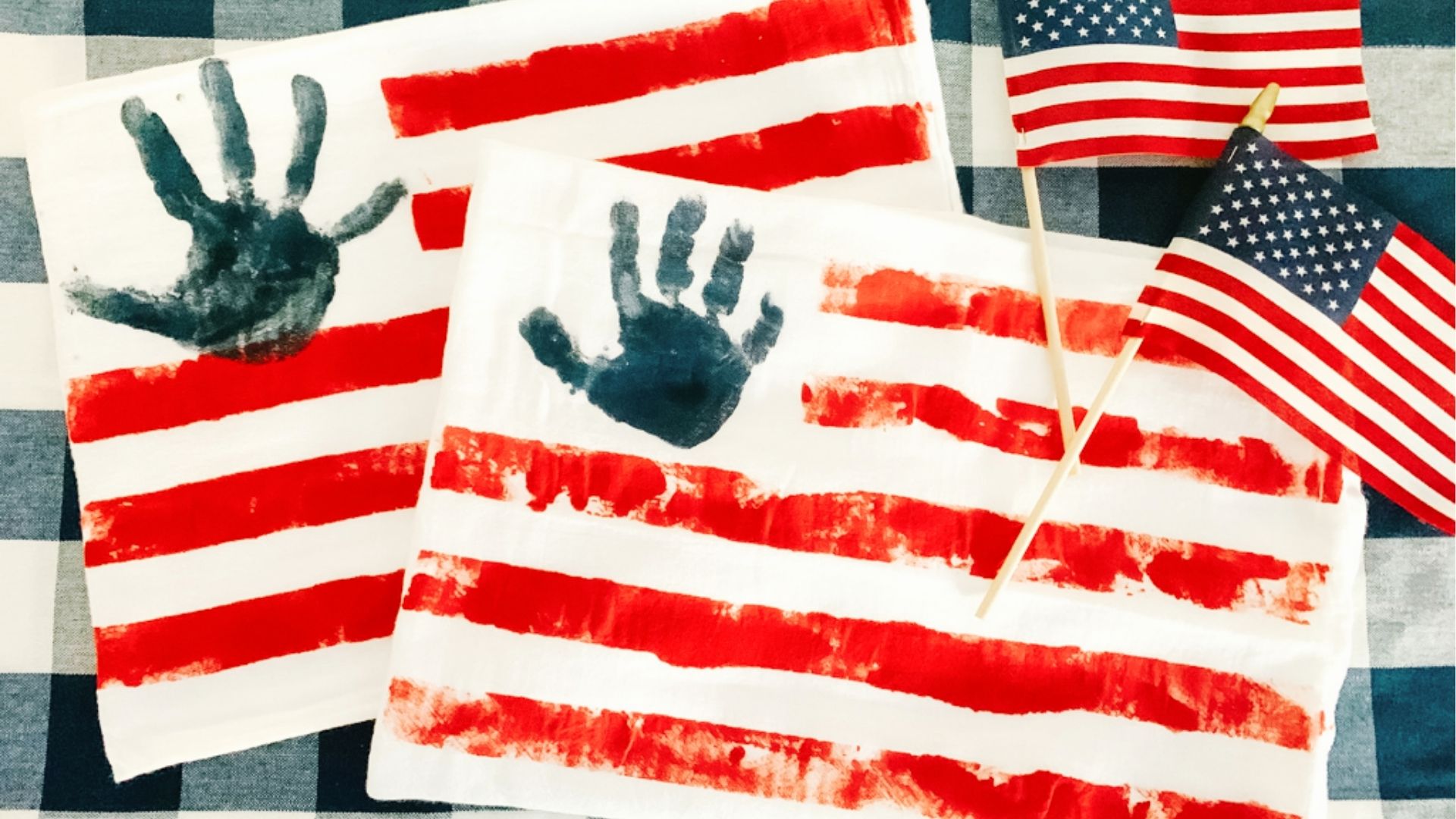 Two handprint American flags on paper with blue painted handprints and red stripes, next to mini flags.