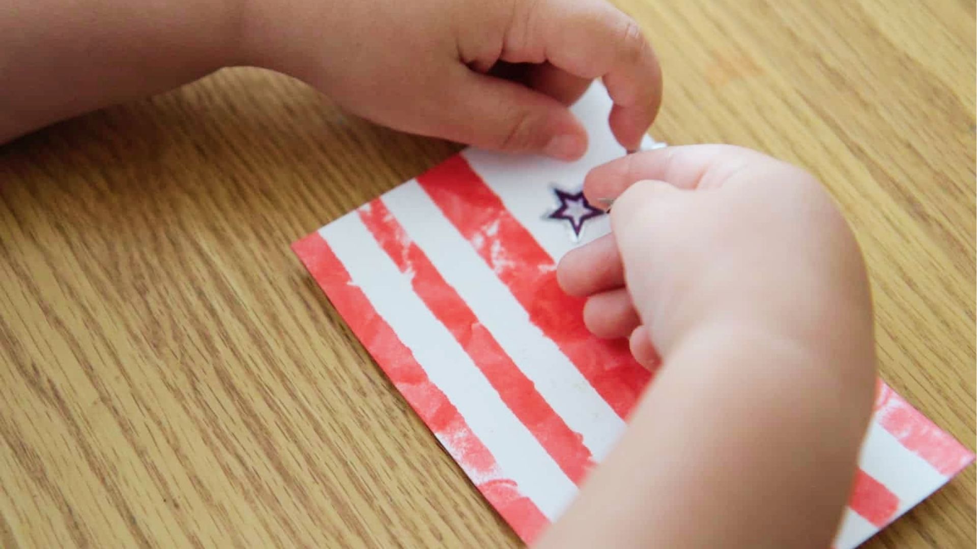 Toddler hands place a purple star sticker onto a hand-painted American flag with red stripes.