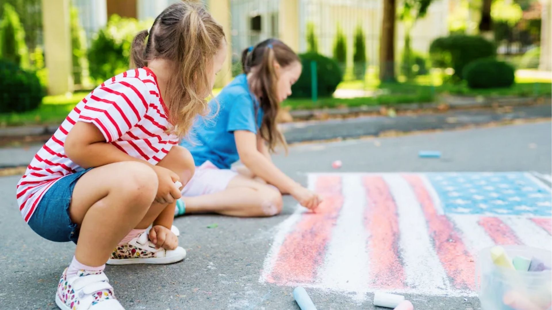 Two toddlers in red stripes and blue shirts crouch to color a large American flag on the sidewalk with chalk.