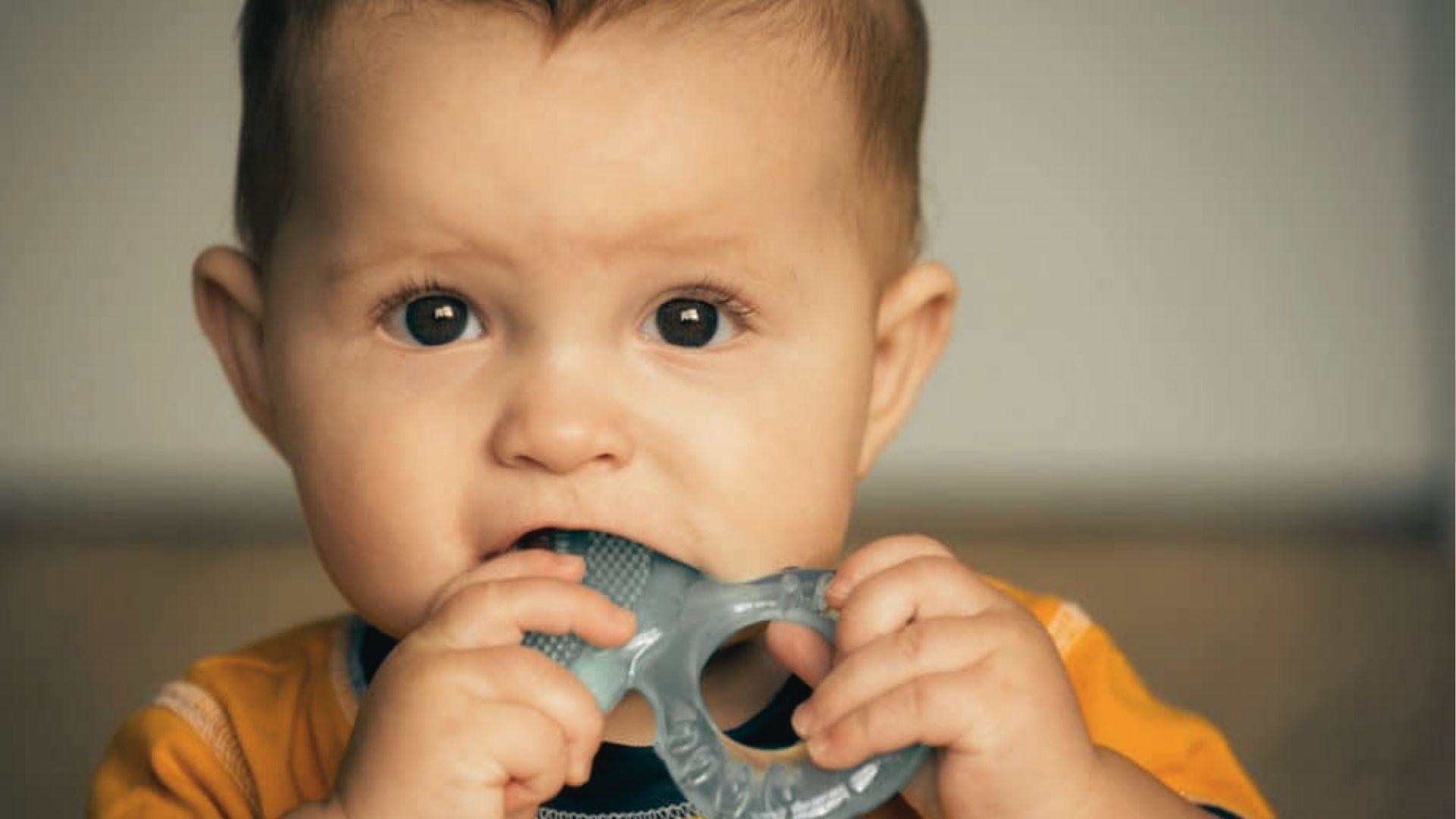 Baby in a mustard yellow outfit chewing on a blue silicone teething ring with both hands.