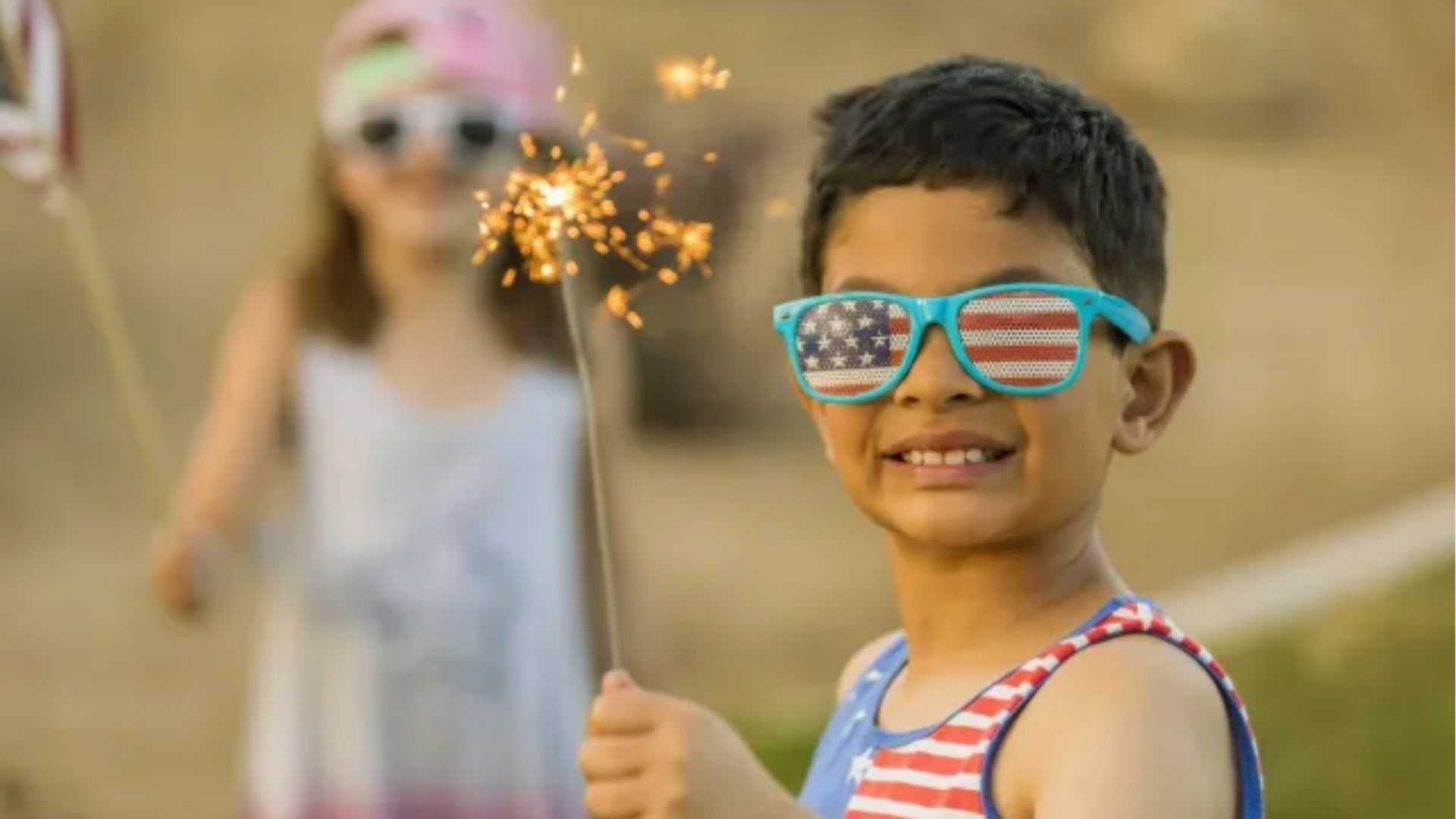 Young boy in American flag sunglasses and tank top holds a lit sparkler, with a girl holding a flag behind him.