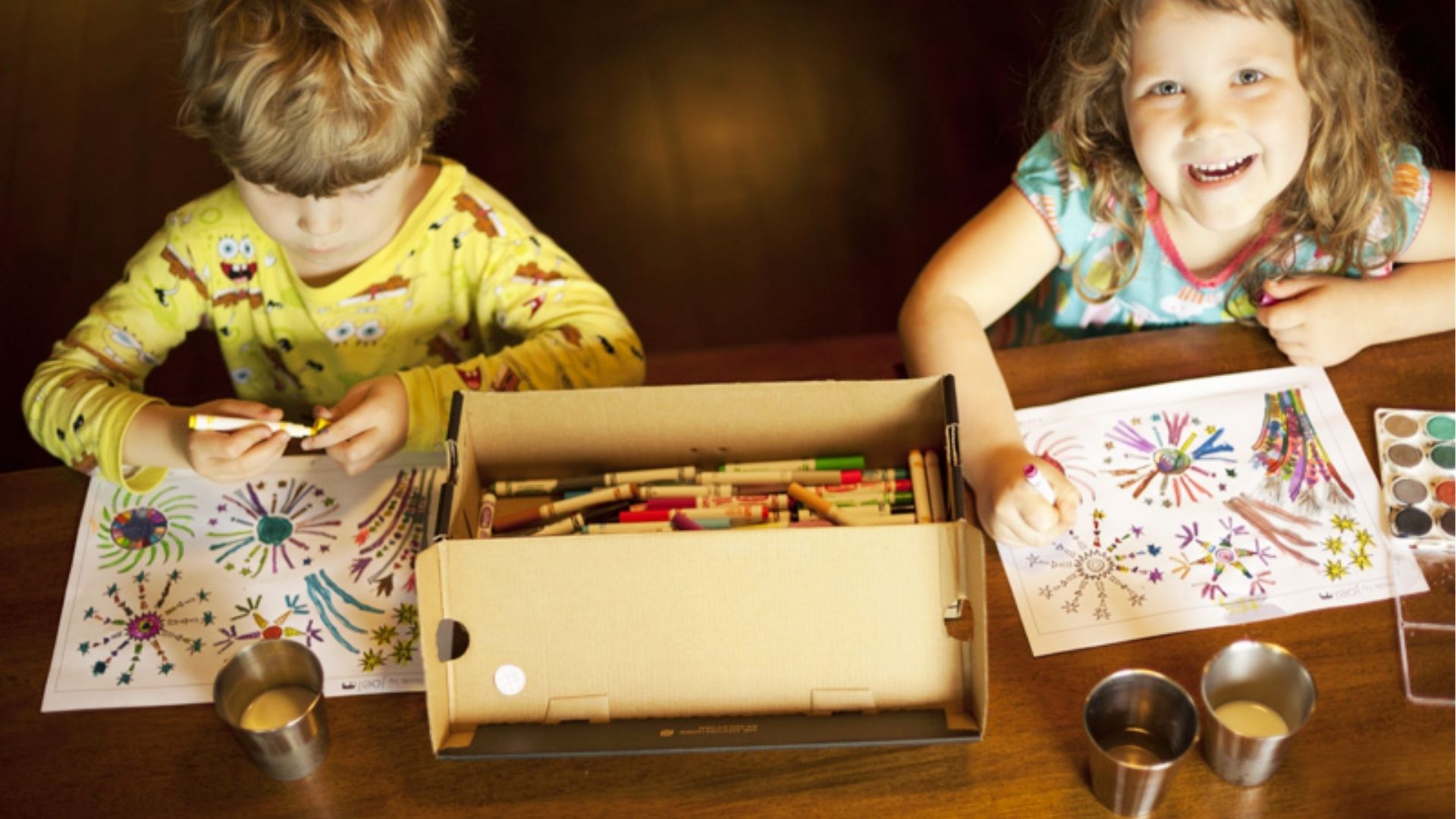 Two children in pajamas color firework-themed pages with markers and crayons at a wooden table.
