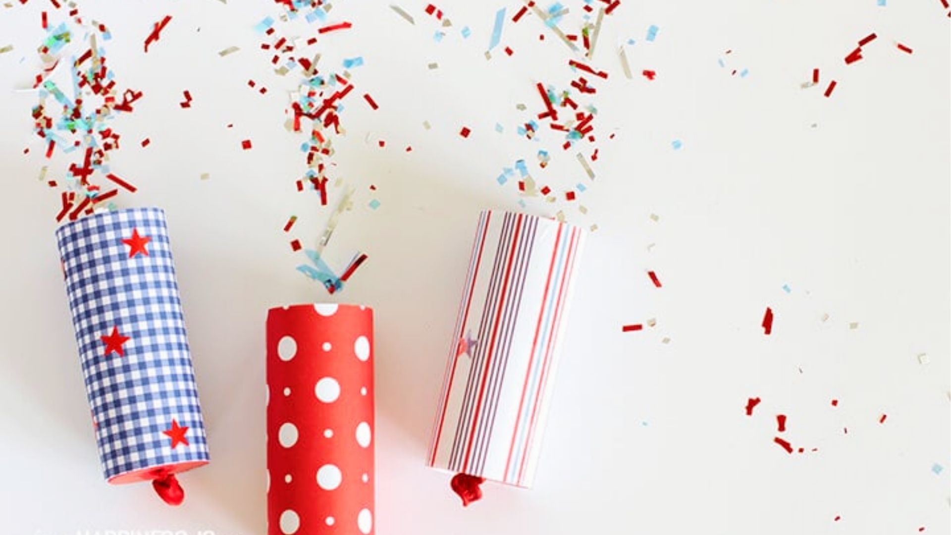 Three DIY confetti poppers made from toilet paper rolls in gingham, polka dot, and stripes, bursting with red and blue confetti.
