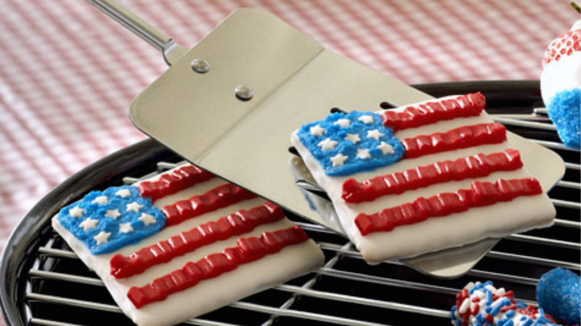 Two rectangular flag cookies with blue stars and red icing stripes rest on a grill spatula over a BBQ